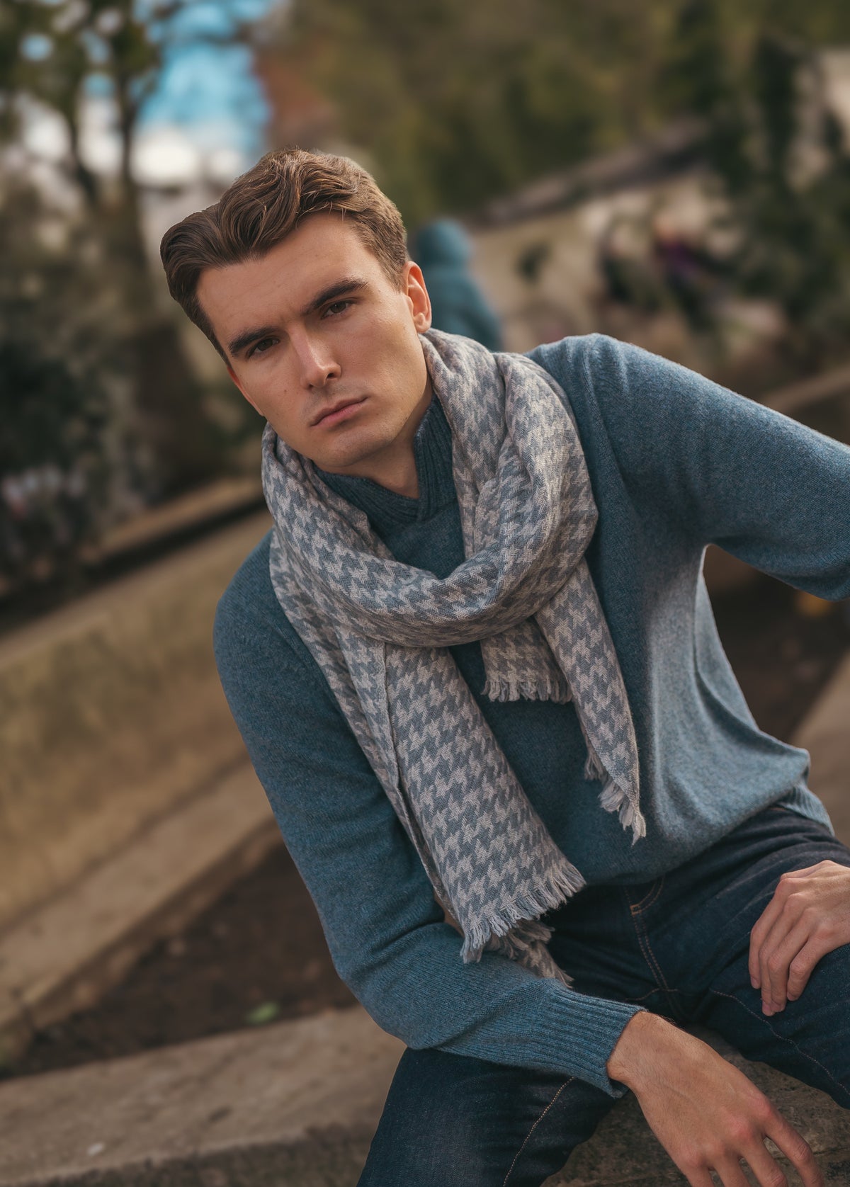 A young man with short brown hair sits outdoors. He wears a blue sweater, dark jeans, and a gray patterned scarf. The background is blurred with greenery and a concrete structure visible.