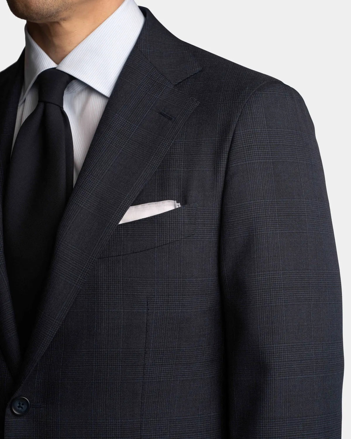 A close-up of a man in the Cavour Navy Check Hightwist Wool Suit, featuring a dark plaid jacket, white pocket square, striped shirt, and black tie. The focus is on his upper torso against a white background—ideal for business travel. , available at Richard Gelding Mayfair