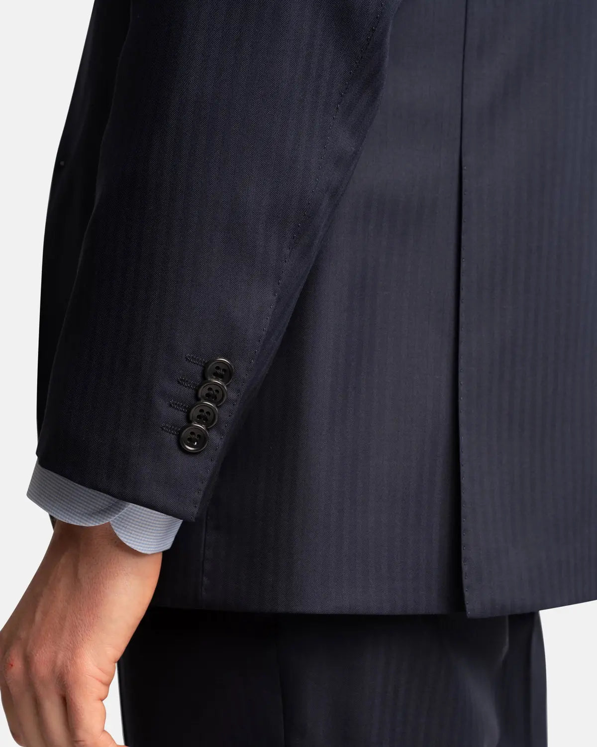 Close-up of a person wearing the Cavour Navy Herringbone Super 150's Wool Suit, highlighting the arm and sleeve with four black cuff buttons. A light blue shirt cuff is visible beneath the jacket as their hand rests at their side. , available at Richard Gelding Mayfair