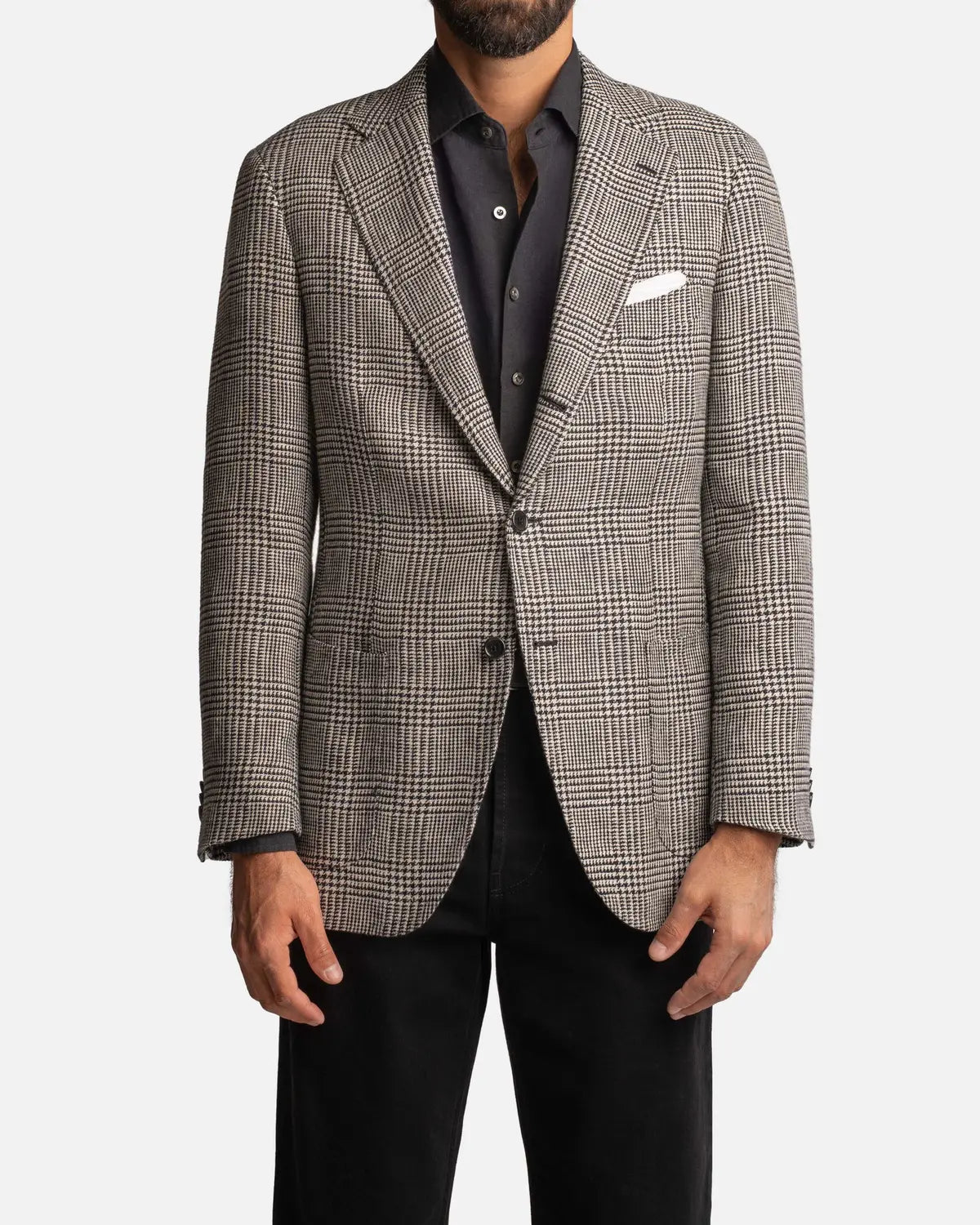 A man wears the Cavour Black/Grey Linen Glencheck Blazer, a single-breasted jacket with a classic notch lapel and two buttons, over a dark shirt with a white pocket square and black pants; only his torso and upper legs are visible. , available at Richard Gelding Mayfair