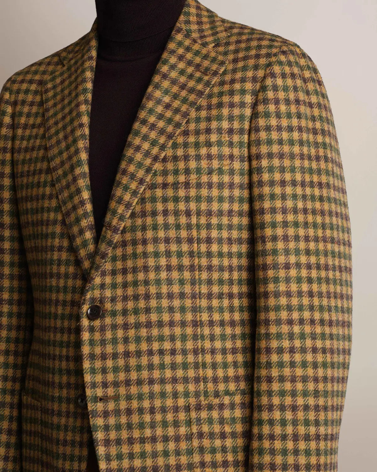 A Brown Shepherds Check Tweed Blazer by Cavour in a brown, green, and tan checkered pattern with notched lapels and two brown buttons, worn over a dark brown turtleneck sweater against a plain, light-colored background. , available at Richard Gelding Mayfair