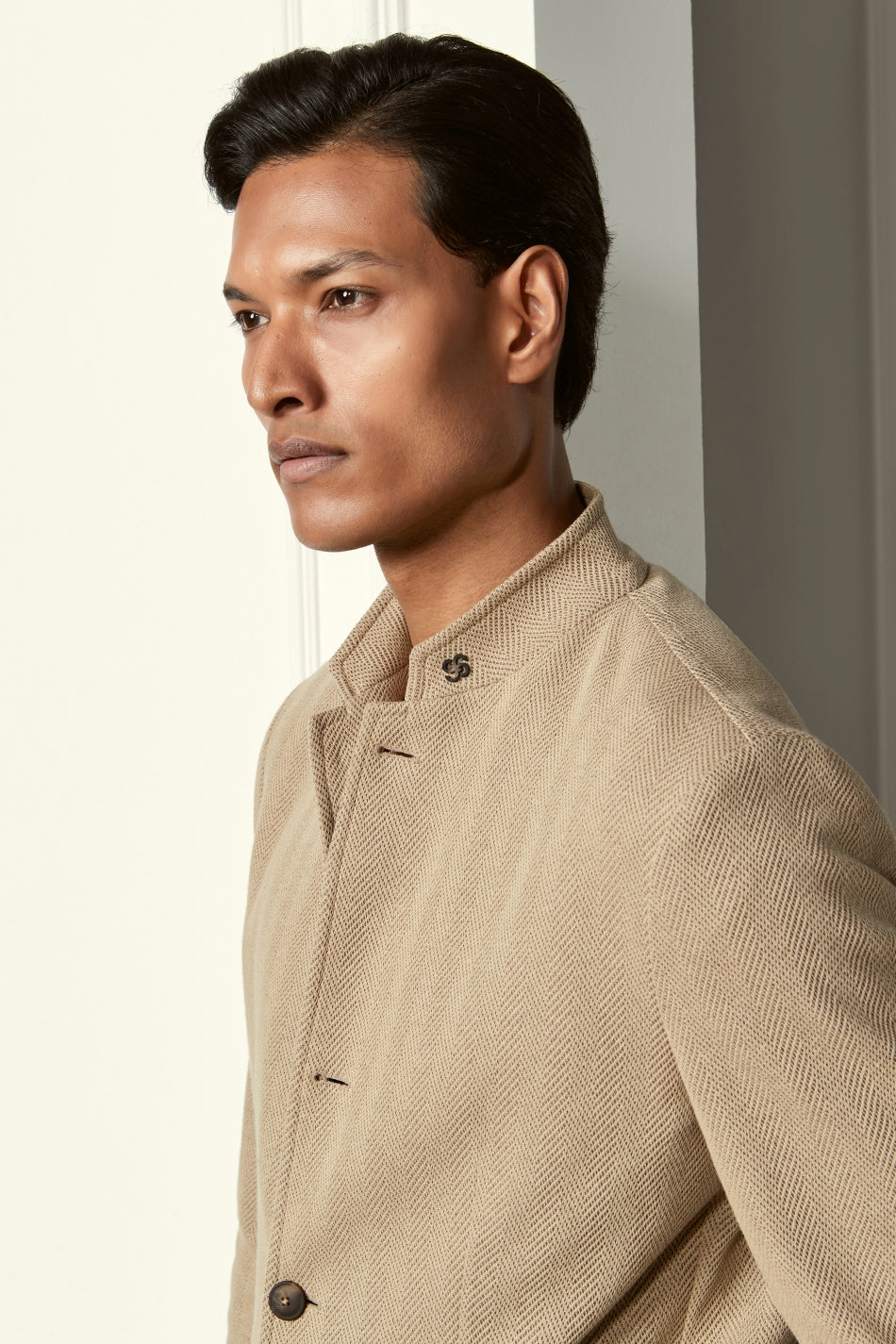 A man with medium brown skin and short, dark hair looks to the side in soft light, wearing the Gran Sasso Beige Herringbone Cotton Blend Jacket with a high collar and decorative buttons against a neutral background. , available at Richard Gelding Mayfair