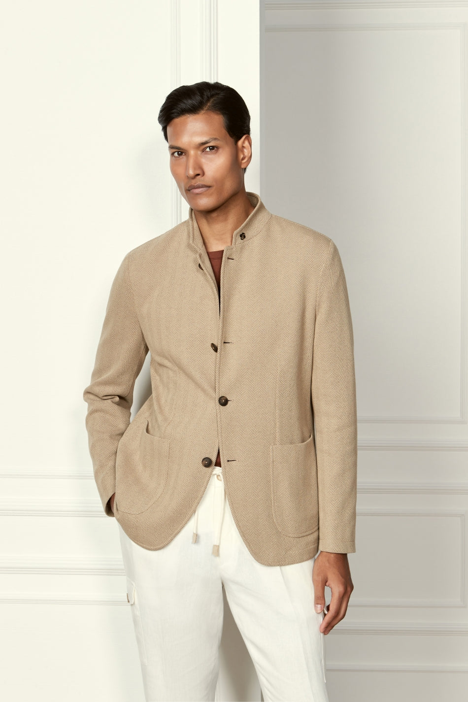 A person with short dark hair and a neutral expression stands indoors, wearing the Gran Sasso Beige Herringbone Cotton Blend Jacket over a brown shirt and white pants. The modern, minimalist style is highlighted by one hand casually in their pocket. , available at Richard Gelding Mayfair