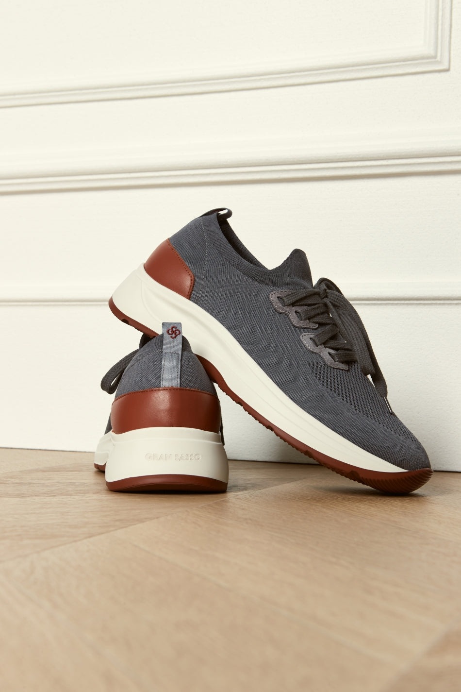 The Gran Sasso Grey Knitted Sneaker is shown indoors on a wooden floor, with one comfortable shoe upright and the other leaning to display the logo on the heel and the textured white sole with brown accents. , available at Richard Gelding Mayfair