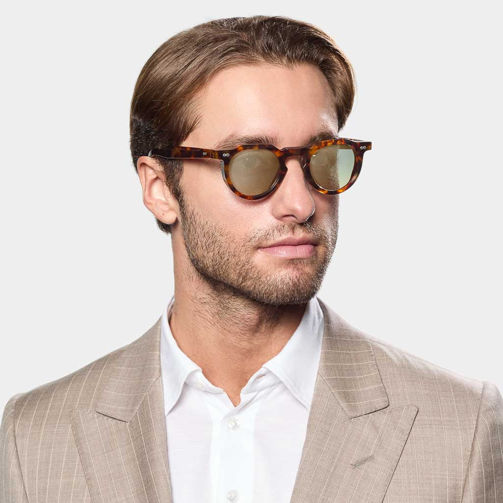 A man with short brown hair and stubble wears TBD Eyewear Hem Eco Spotted Havana | Mirror Green sunglasses and a beige pinstripe suit over a white collared shirt, facing slightly right against a plain light background. , available at Richard Gelding Mayfair