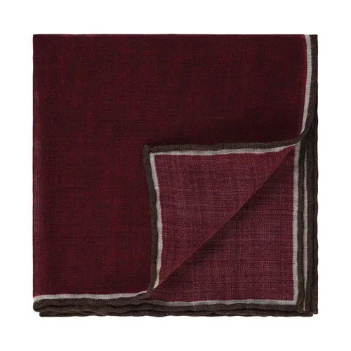 A folded Amanda Christensen wool pocket square with hand rolled edges is displayed on a white background. The printed burgundy fabric features a thin gray and black border, with the bottom right corner turned up to reveal the reverse side and border detailing. , available at Richard Gelding Mayfair