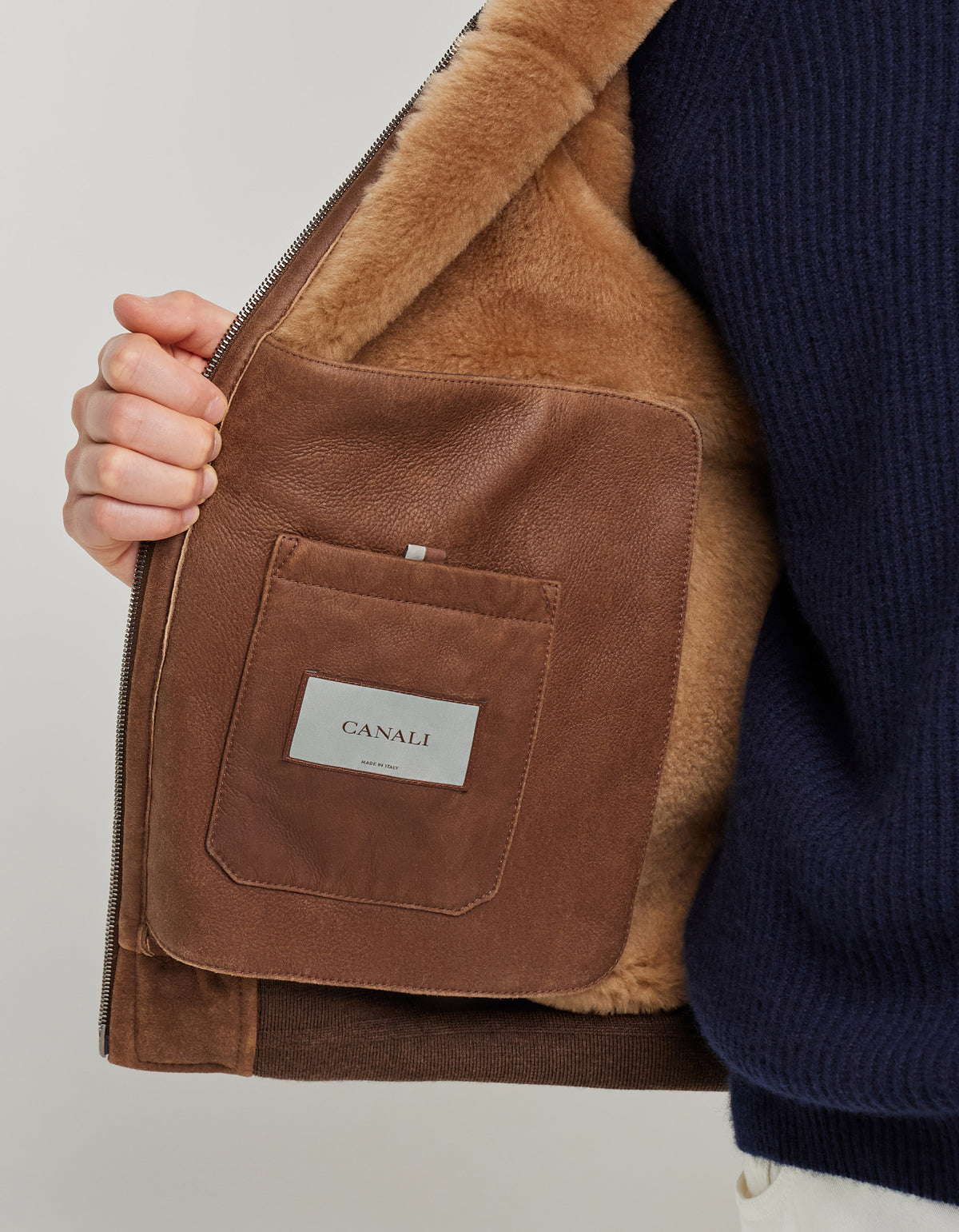 A person in a navy sweater holds open a Canali Brown Merino Shearling Lambskin Suede Blouson, revealing a brown leather interior pocket with a rectangular CANALI label stitched on it. The pocket and the soft lambskin suede-like lining stand out against the jacket’s outer fabric. , available at Richard Gelding Mayfair