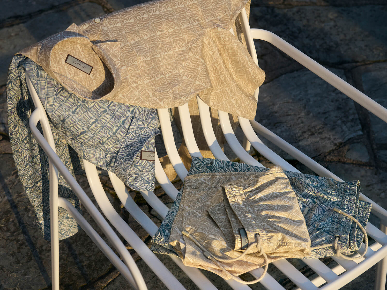 A white metal lounge chair displays Canali Blue Printed Swim Shorts, folded in warm sunlight on a stone-tiled surface. The shorts feature subtle geometric prints and branded tags in soft blue tones. , available at Richard Gelding Mayfair