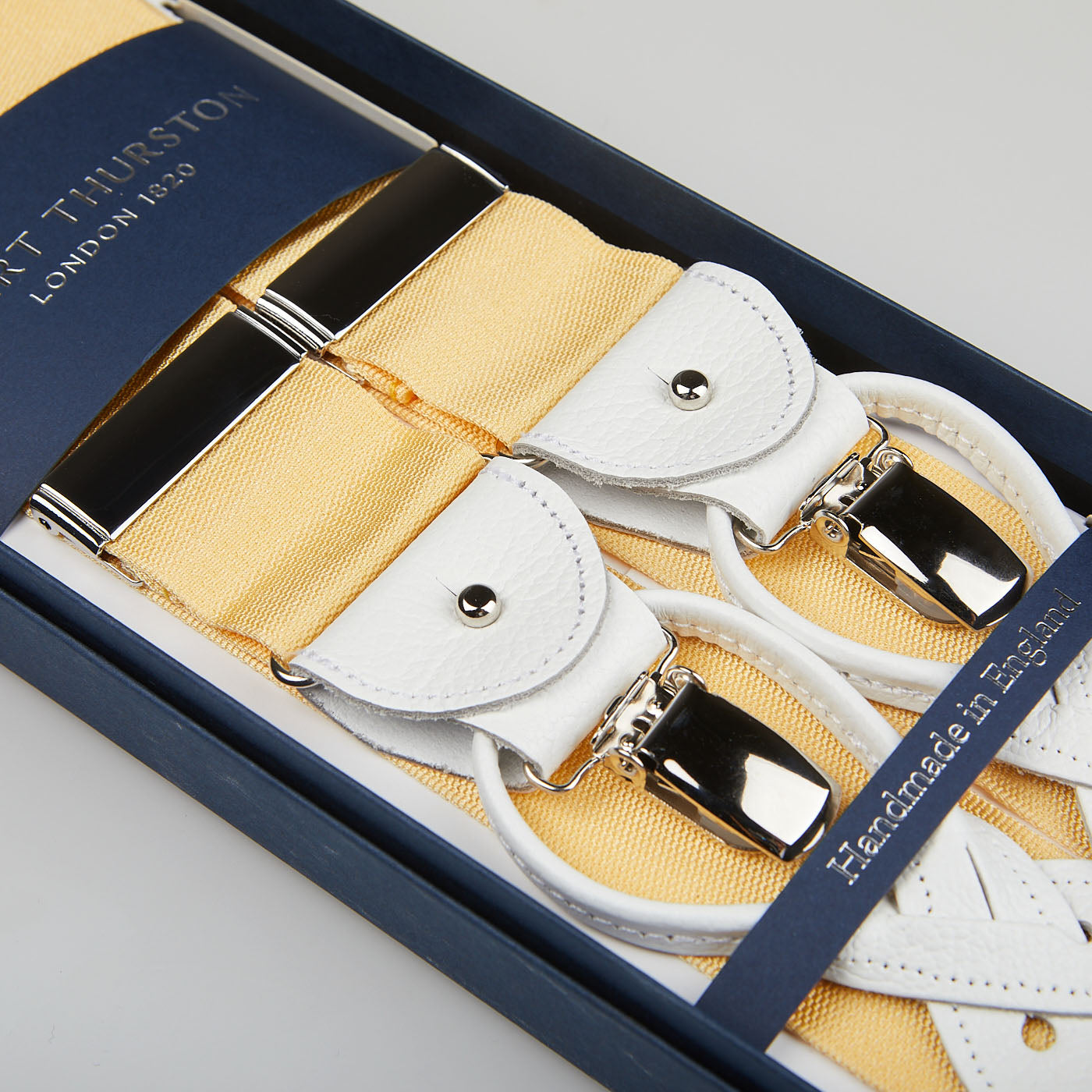 A box of Champagne Nylon Leather 40mm Braces with white leather and metal clips reads, "Handmade in England by Albert Thurston," emphasizing the excellence and tradition of this esteemed British brand.