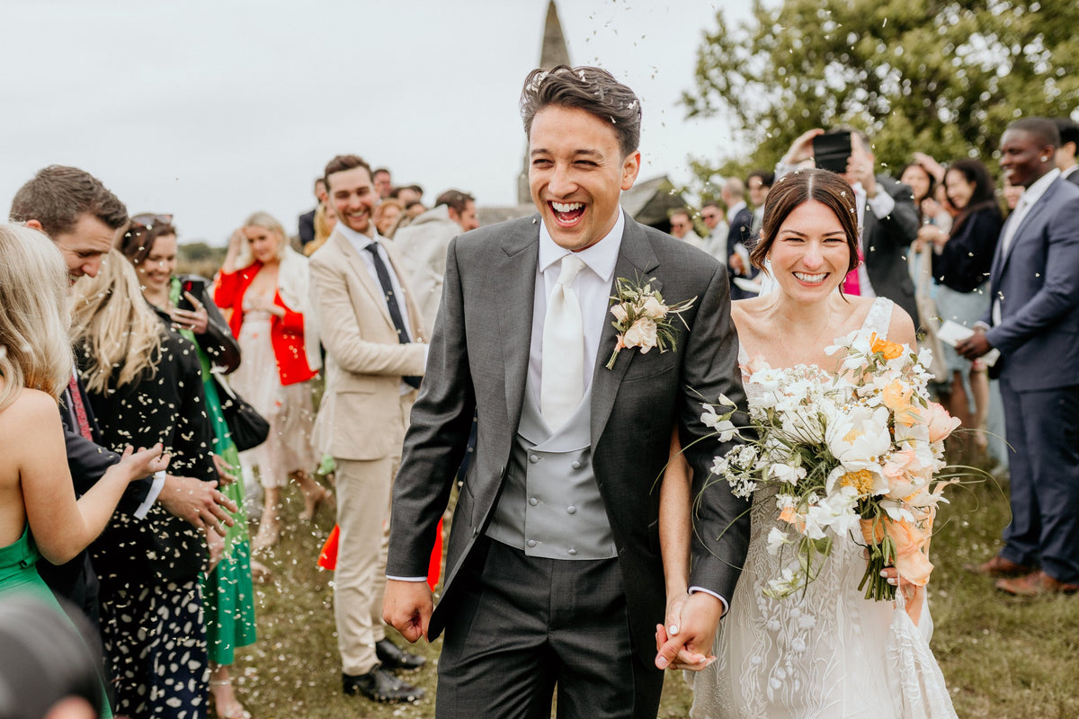 A joyful bride and groom walk hand in hand outdoors, smiling as guests throw confetti. The groom wears a gray suit and the bride holds a bouquet of white and peach flowers. People in festive attire celebrate around them on a grassy area. , available at Richard Gelding Mayfair