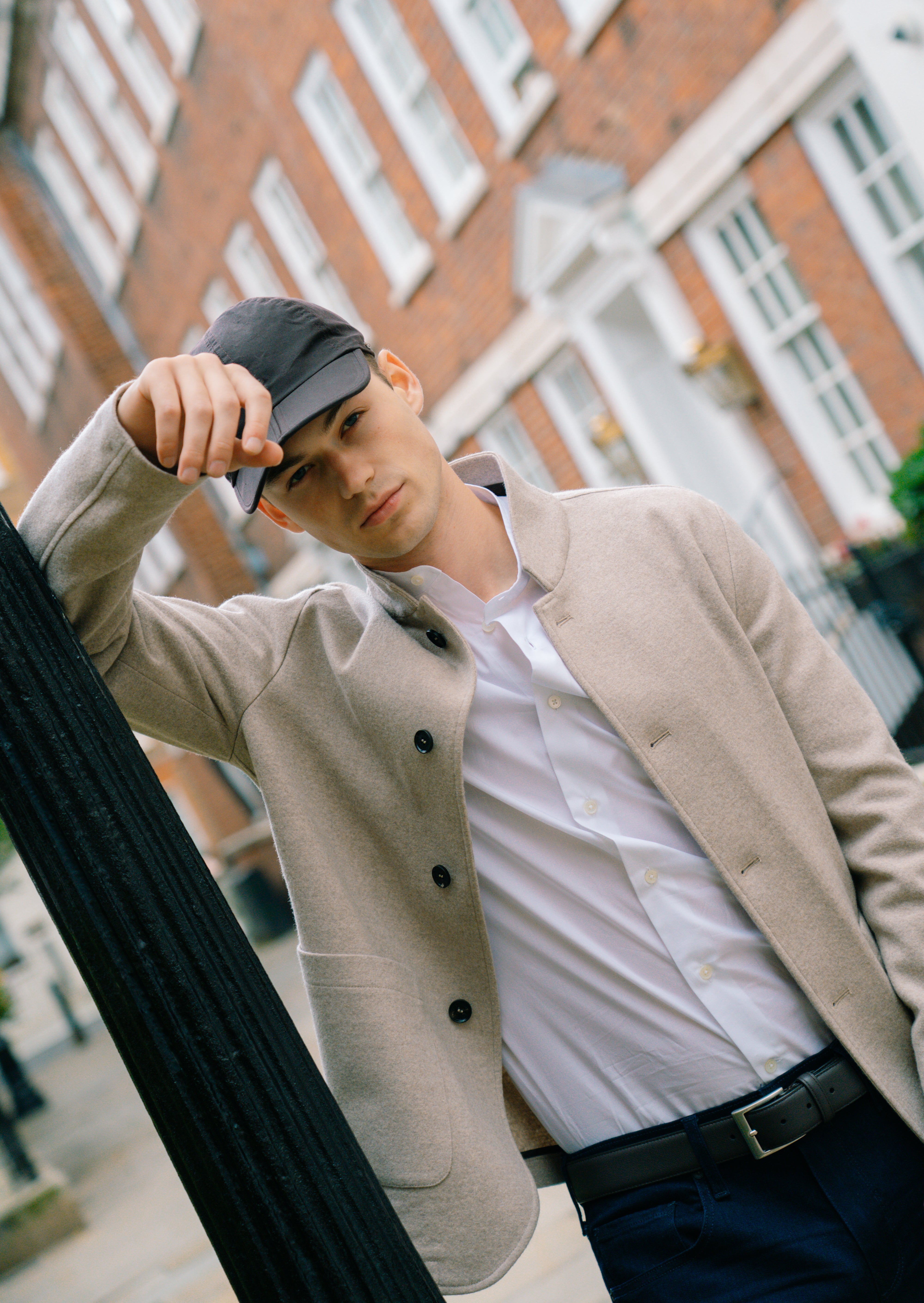 A young man in a beige jacket and Coconut Residence's White Poplin Mandarin Collar Shirt leans against a black lamppost on a city street with red brick buildings and white-trimmed windows, looking at the camera with relaxed confidence. , available at Richard Gelding Mayfair
