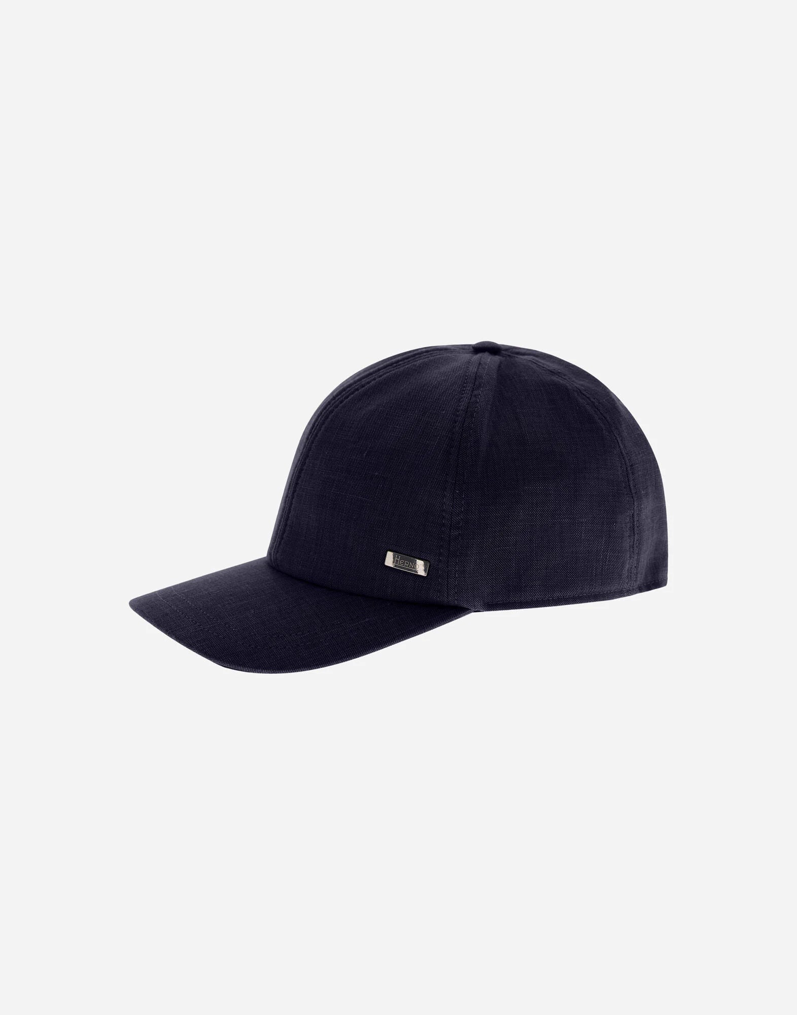 The Herno Navy Wool/Silk/Linen Loro Piana Woven Cap features a dark navy hue, curved brim, rectangular metallic logo on the left panel, visible stitching, and a smooth, structured shape on a light grey background. , available at Richard Gelding Mayfair