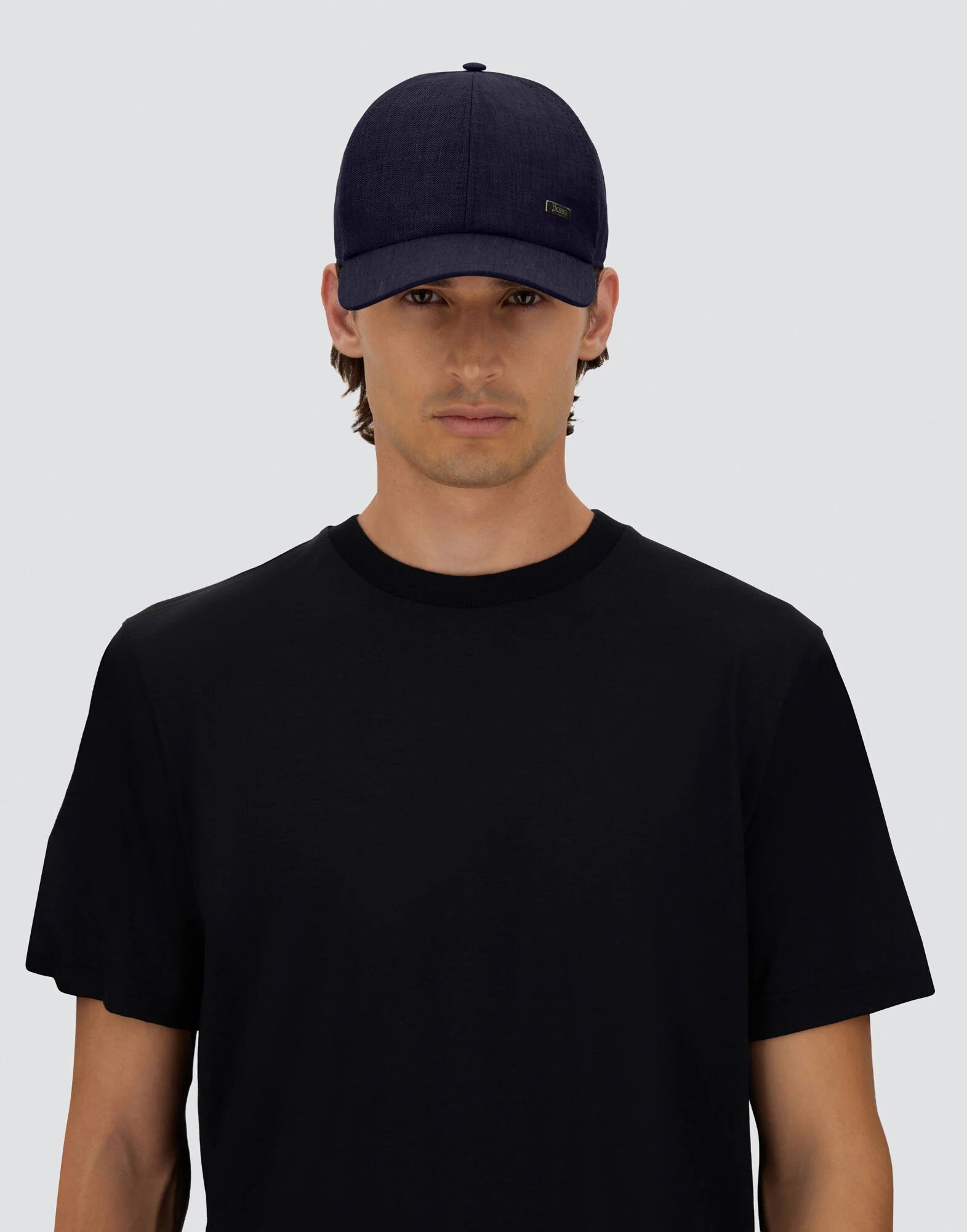 A person wears a Herno Navy Wool/Silk/Linen Loro Piana Woven Cap and a plain black t-shirt, standing against a light gray background with a neutral expression. , available at Richard Gelding Mayfair