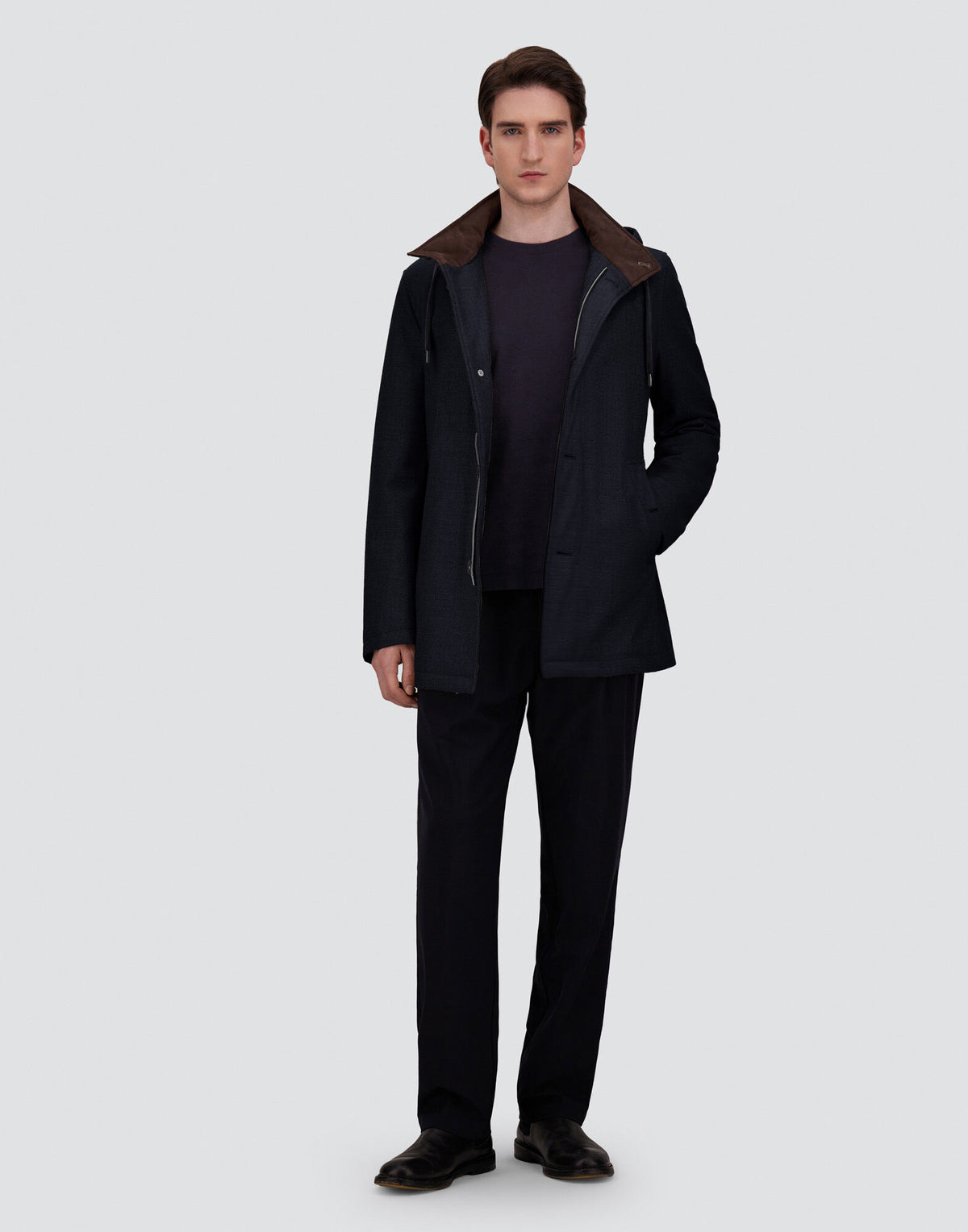 A man stands facing forward against a plain gray background, wearing a navy blue Herno parka crafted from luxurious Loro Piana wool and silk with a removable hood, paired with a dark shirt, dark trousers, and black shoes. His left hand is in his pocket and he has a neutral expression. , available at Richard Gelding Mayfair