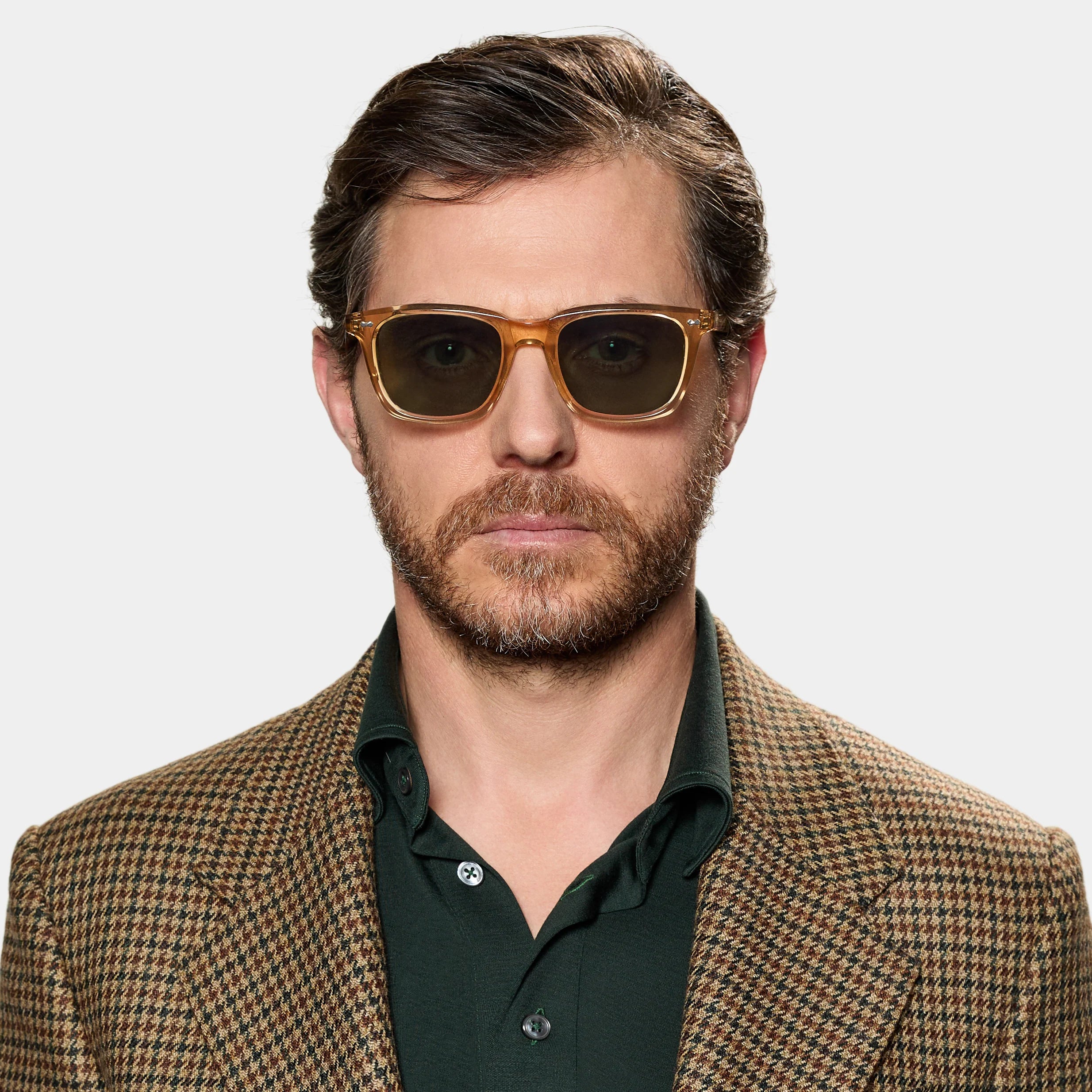 A man with short brown hair, a beard, and mustache wears TBD Eyewear's Carré Eco Champagne | Bottle Green sunglasses. He looks ahead with a neutral expression, dressed in a green collared shirt and brown houndstooth blazer against a white background. , available at Richard Gelding Mayfair
