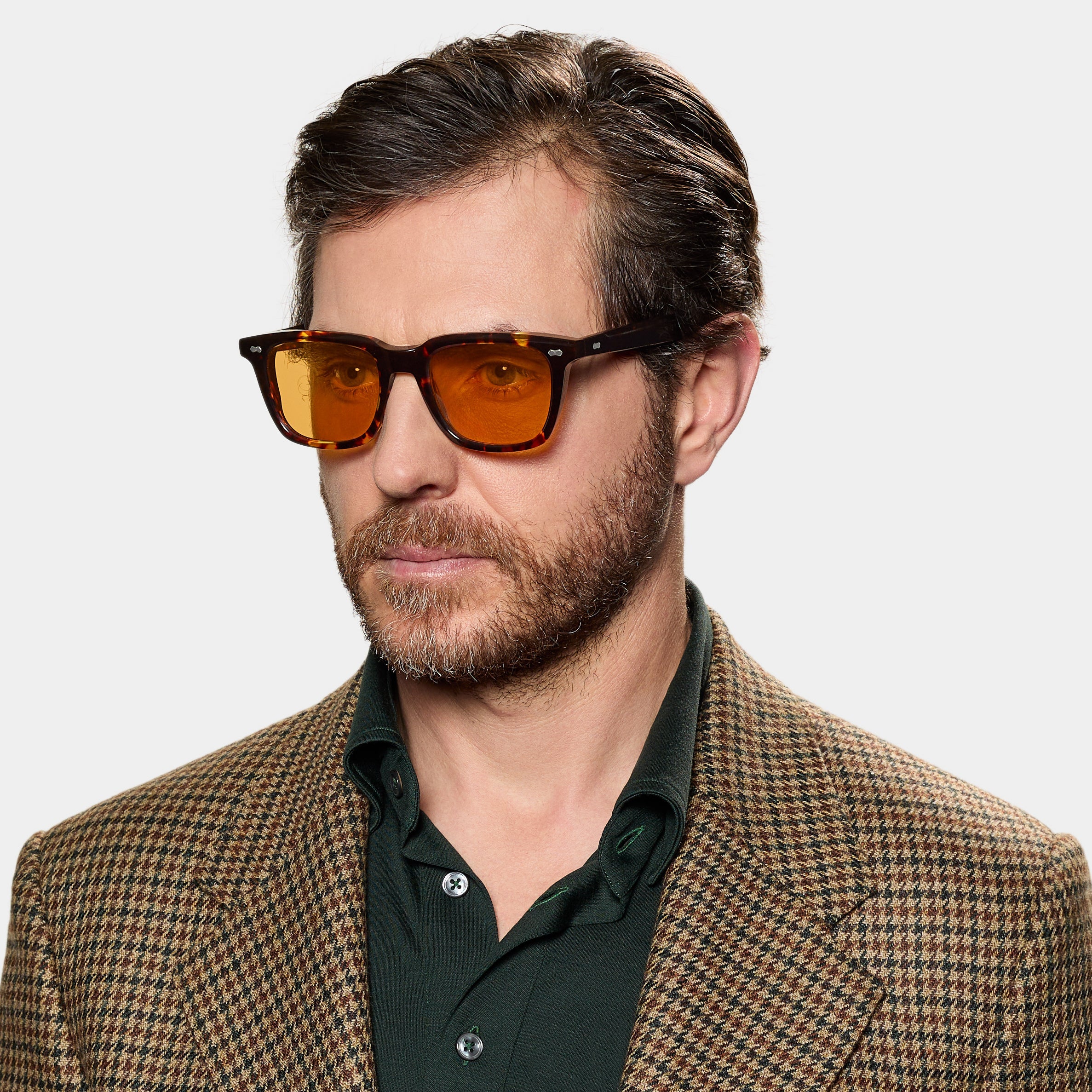 A man with short brown hair and a beard wears TBD Eyewear's Carré Eco Dark Havana | Orange sunglasses, a green collared shirt, and a brown plaid blazer, looking slightly to the side against a plain white background. , available at Richard Gelding Mayfair