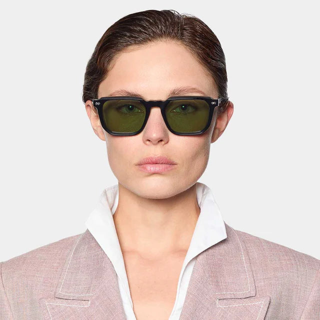 A person with short brown hair wears TBD Eyewear Dart Black | Green sustainable acetate sunglasses handmade in Italy, featuring green mineral glass lenses, paired with a light pink jacket and white shirt, looking ahead against a plain background. , available at Richard Gelding Mayfair