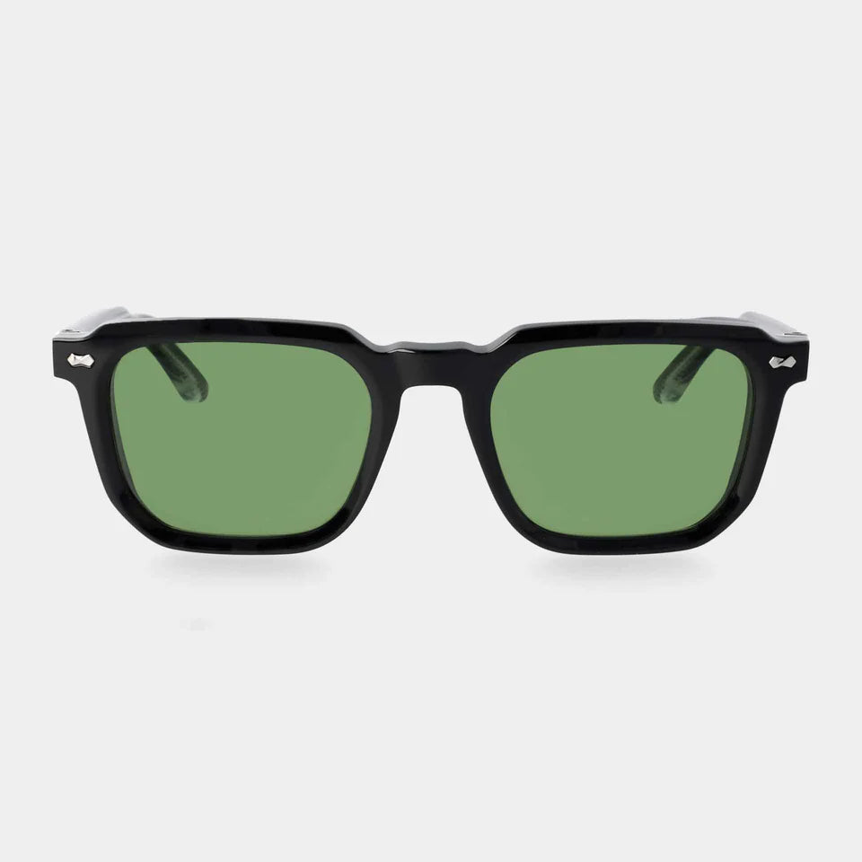 The Dart Black | Green sunglasses by TBD Eyewear feature thick, glossy black sustainable acetate frames and green mineral glass lenses. Handcrafted in Italy, they are shown front-facing on a light gray background with detailed silver hinges at the temples. , available at Richard Gelding Mayfair