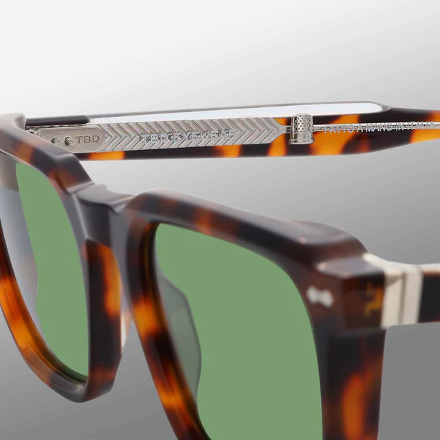 Close-up of TBD Eyewear’s Dart Brown Havana | Green sunglasses, showing green mineral glass lenses and a metal hinge with herringbone texture and engraved details, set against a smooth white-to-light gray gradient background. , available at Richard Gelding Mayfair