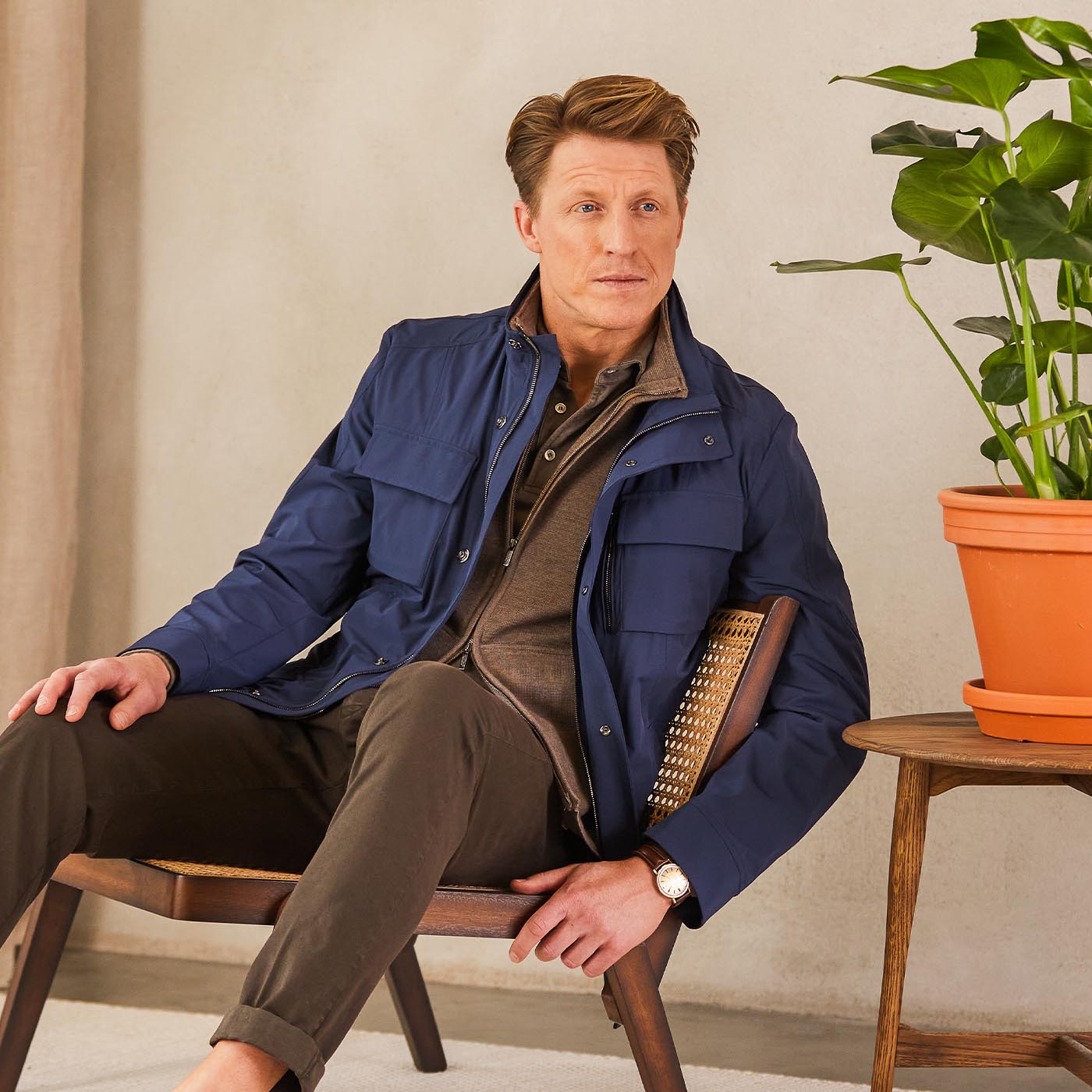 A man wearing a Moorer Dark Blue Lightweight Nylon Field Jacket sits on a chair next to a potted plant.