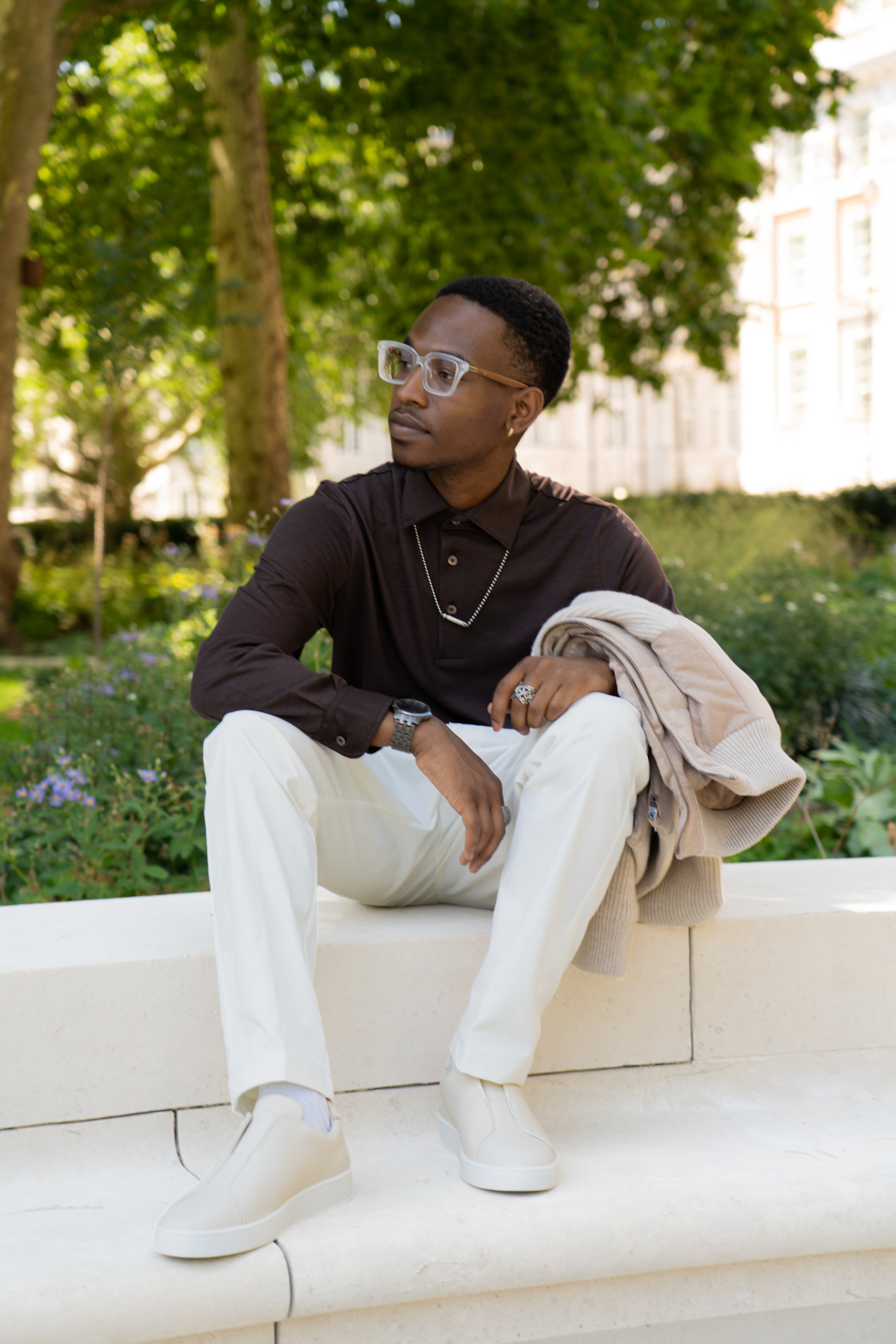 A man sits outdoors on a white stone bench, wearing the Herno Brown Wool Jersey Long Sleeve Polo, paired with white pants and white slip-on shoes. He holds a beige sweater in one hand and looks to the side, sporting clear-framed glasses amid greenery and trees. , available at Richard Gelding Mayfair