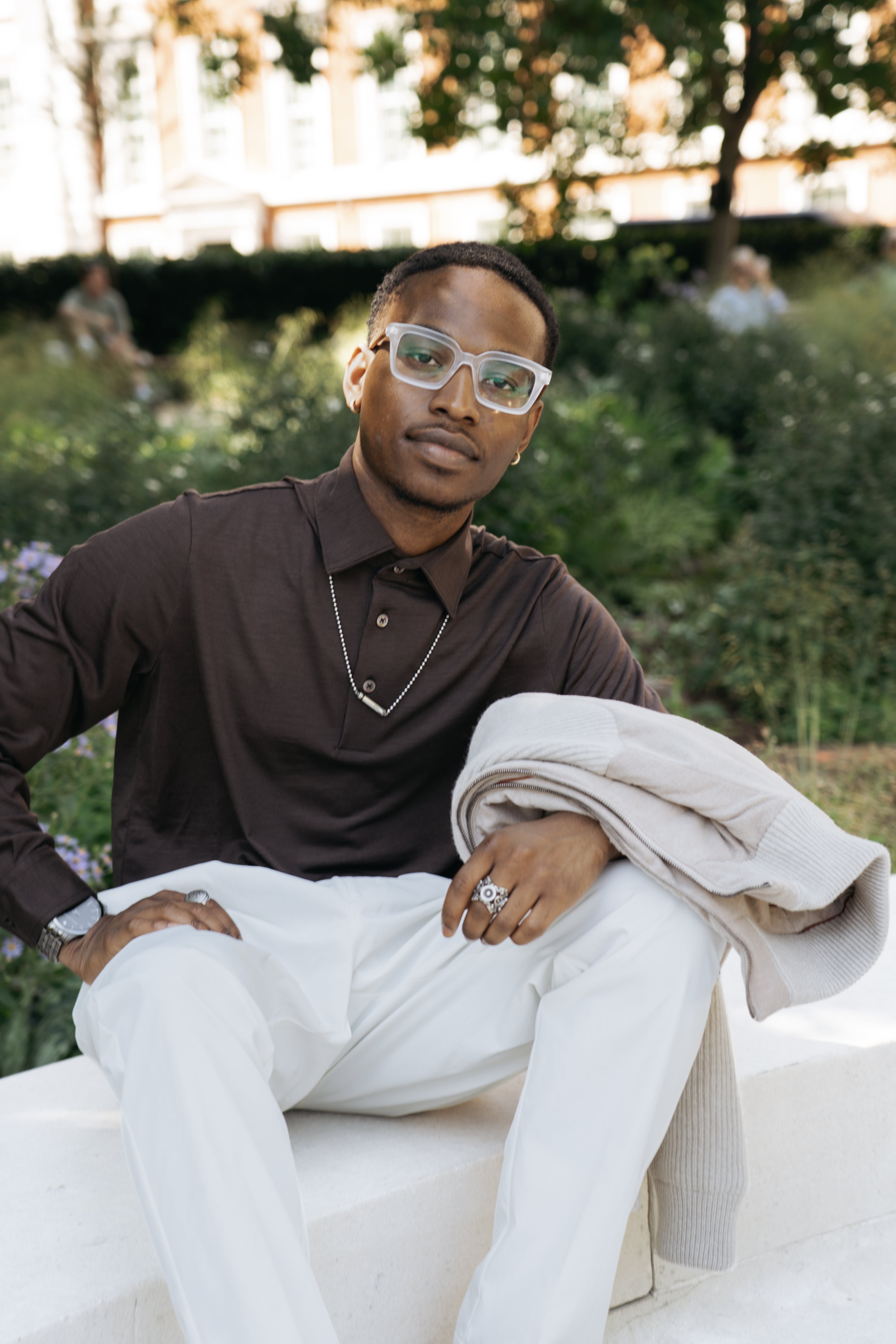 A man with short hair sits outdoors on a white bench, wearing clear glasses, white pants, jewelry, and the Herno Brown Wool Jersey Long Sleeve Polo. He holds a beige sweater in his lap, with green shrubs and trees in the background. , available at Richard Gelding Mayfair