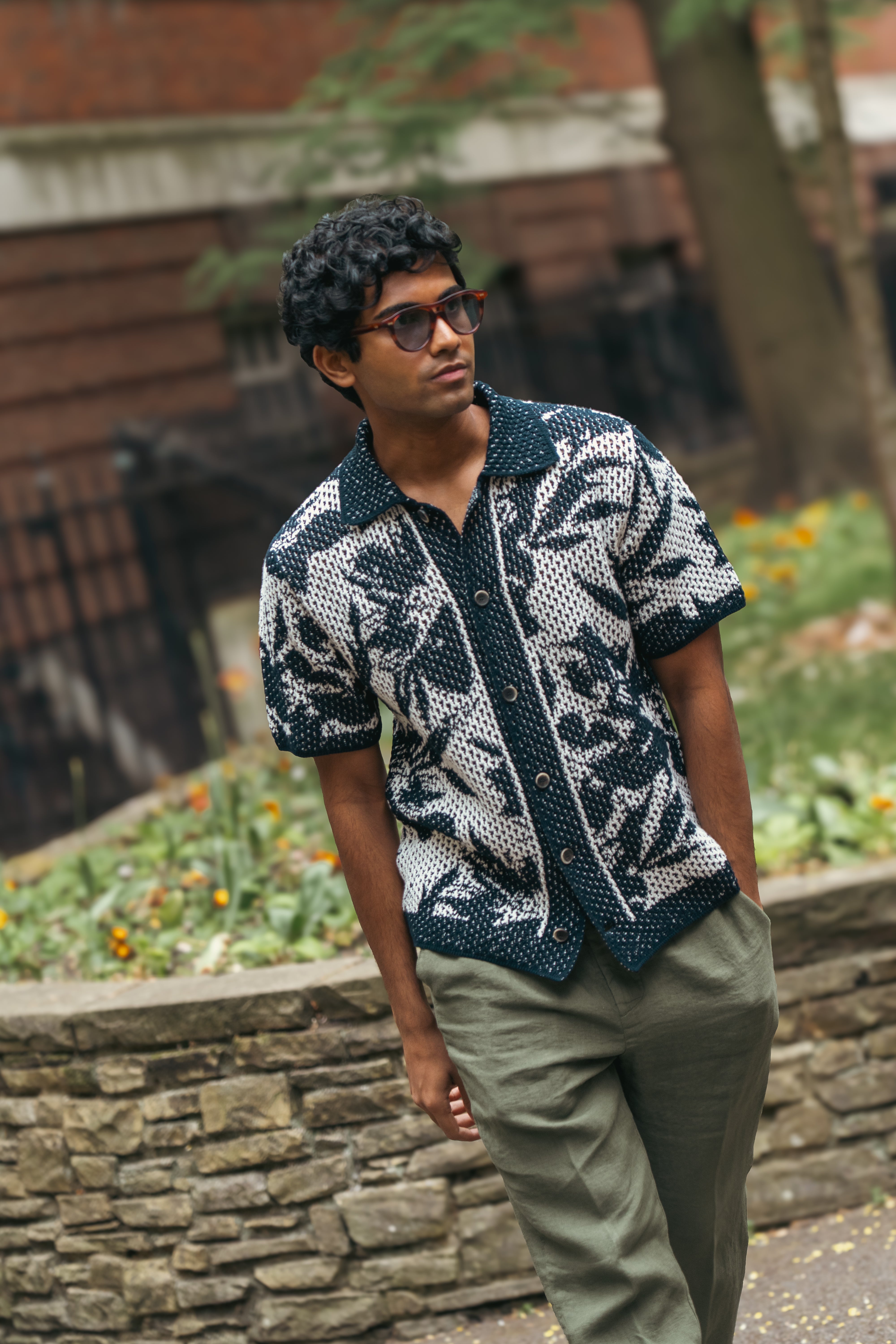 A man with curly hair and sunglasses stands by a stone wall and garden, wearing the Altea Navy Cream Cotton Flower Open Weave short-sleeve shirt, paired with olive pants. He looks off to the side; trees and a brick building are in the background. , available at Richard Gelding Mayfair