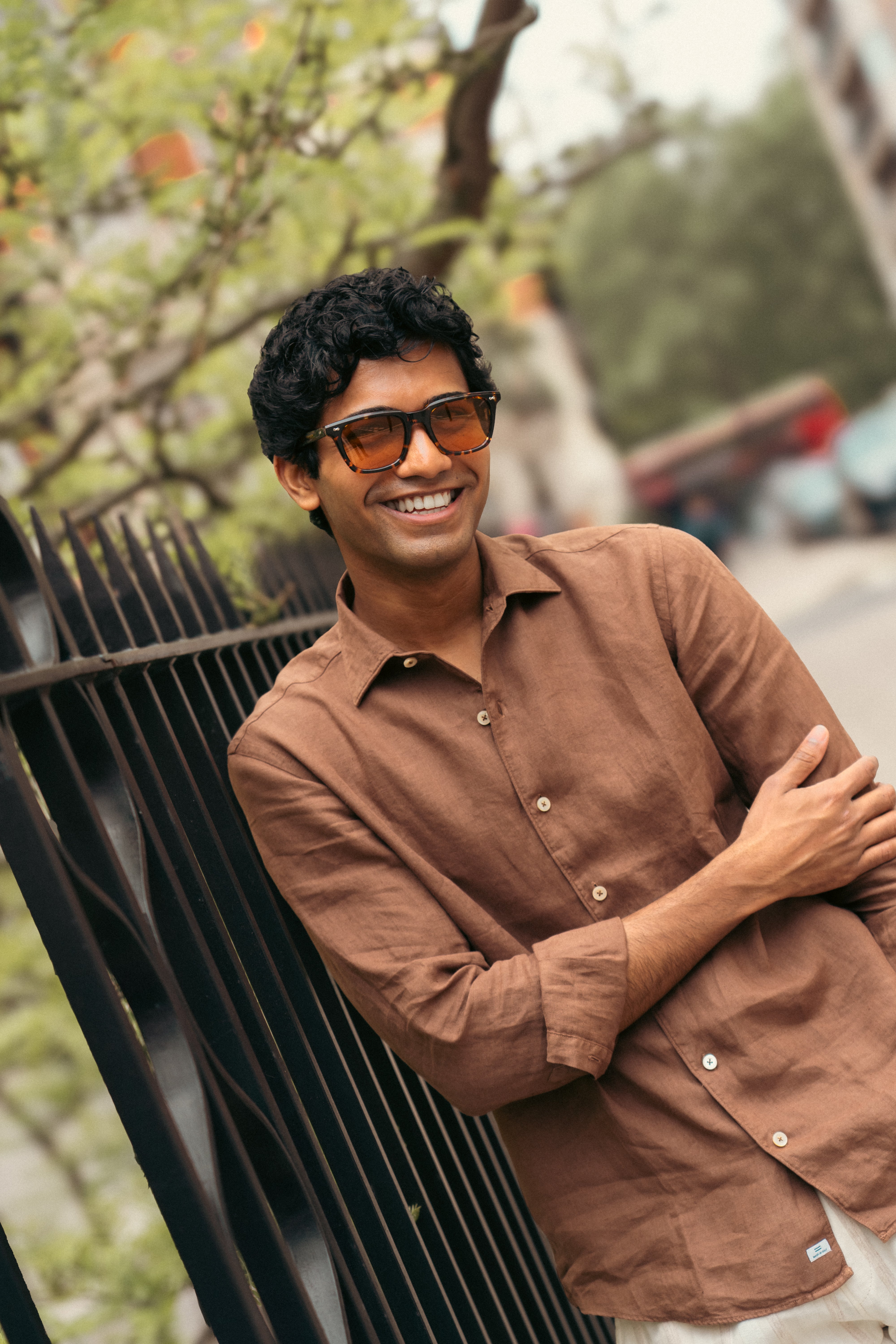 A smiling man with dark curly hair and sunglasses stands outdoors, leaning casually against a black fence. He wears the Alessandro Gherardi Tobacco Brown Linen One Piece Collar Casual Shirt with white pants. Trees and a city street blur behind him. , available at Richard Gelding Mayfair