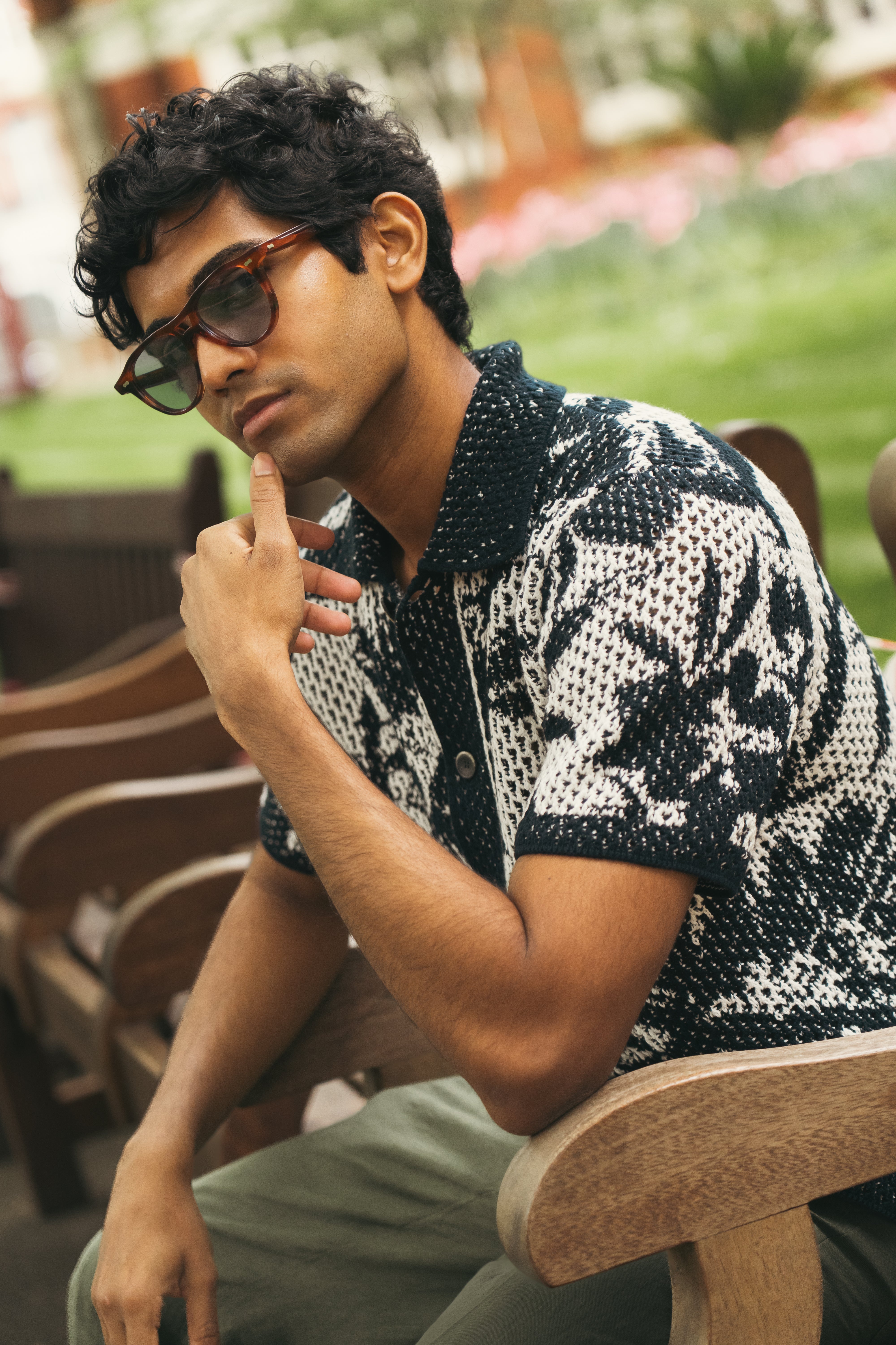 A man with short, curly dark hair and brown skin sits on a wooden bench outdoors, thoughtfully wearing Altea's Navy Cream Cotton Flower Open Weave shirt and tortoiseshell sunglasses, paired with olive-green pants. , available at Richard Gelding Mayfair