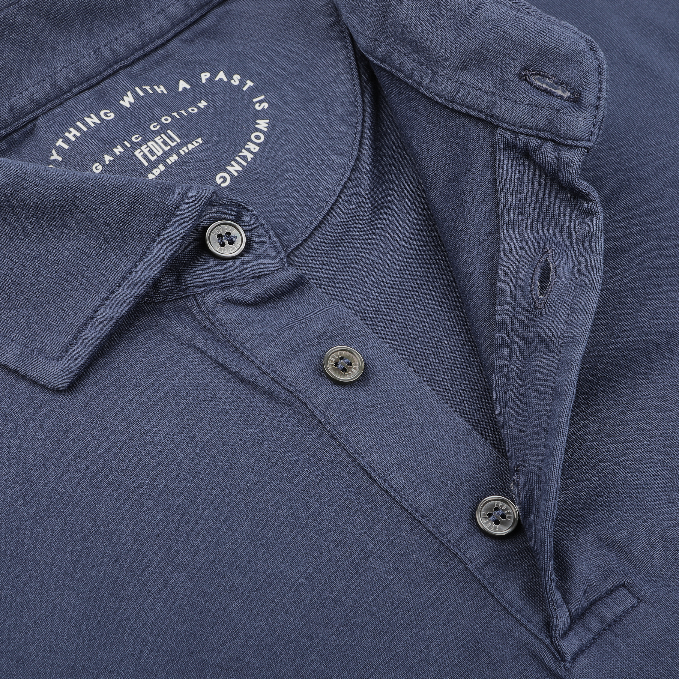 A close-up of the Fedeli Dark Blue Organic Cotton LS Polo Shirt showcases undone buttons, revealing a logo and text on the inside collar. Made from organic Giza cotton, this elegant long-sleeved polo offers sophistication and comfort.