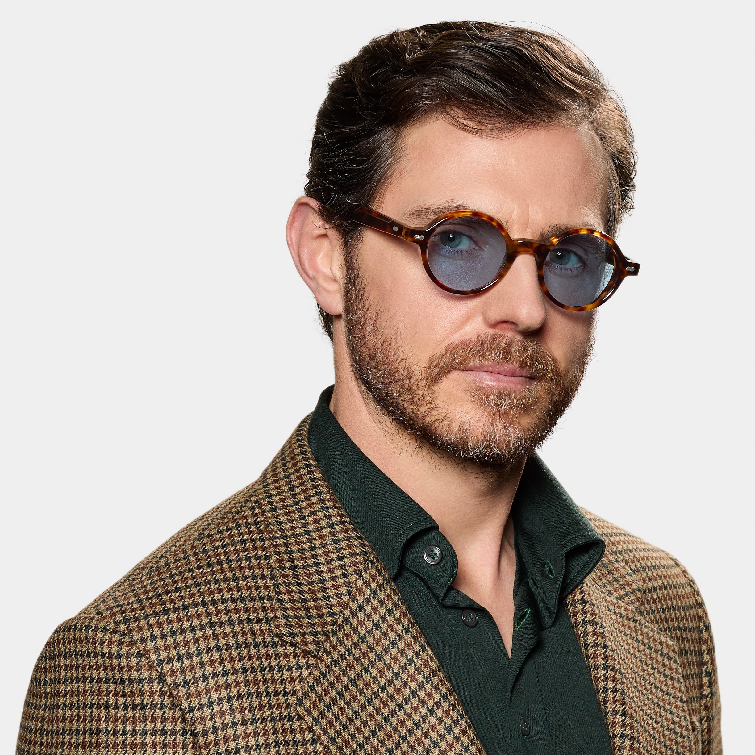 A man with short brown hair and a beard wears TBD Eyewear's Giotto Eco Spotted Havana | Blue round sunglasses, a brown and tan houndstooth blazer, and a dark green shirt. He poses neutrally against a plain white background. , available at Richard Gelding Mayfair