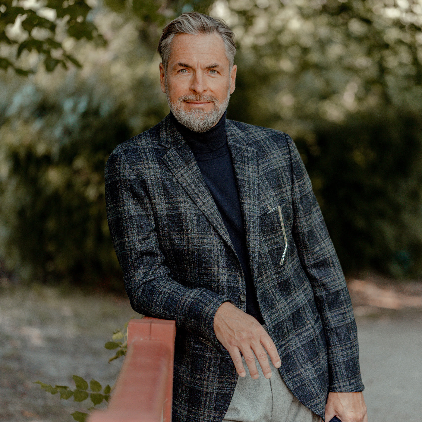 A middle-aged man with gray hair and a beard stands outdoors by a red railing, wearing a Gran Sasso Navy Blue Extra Fine Merino Wool Rollneck under a slim fit plaid blazer and light pants. Trees fill the background.