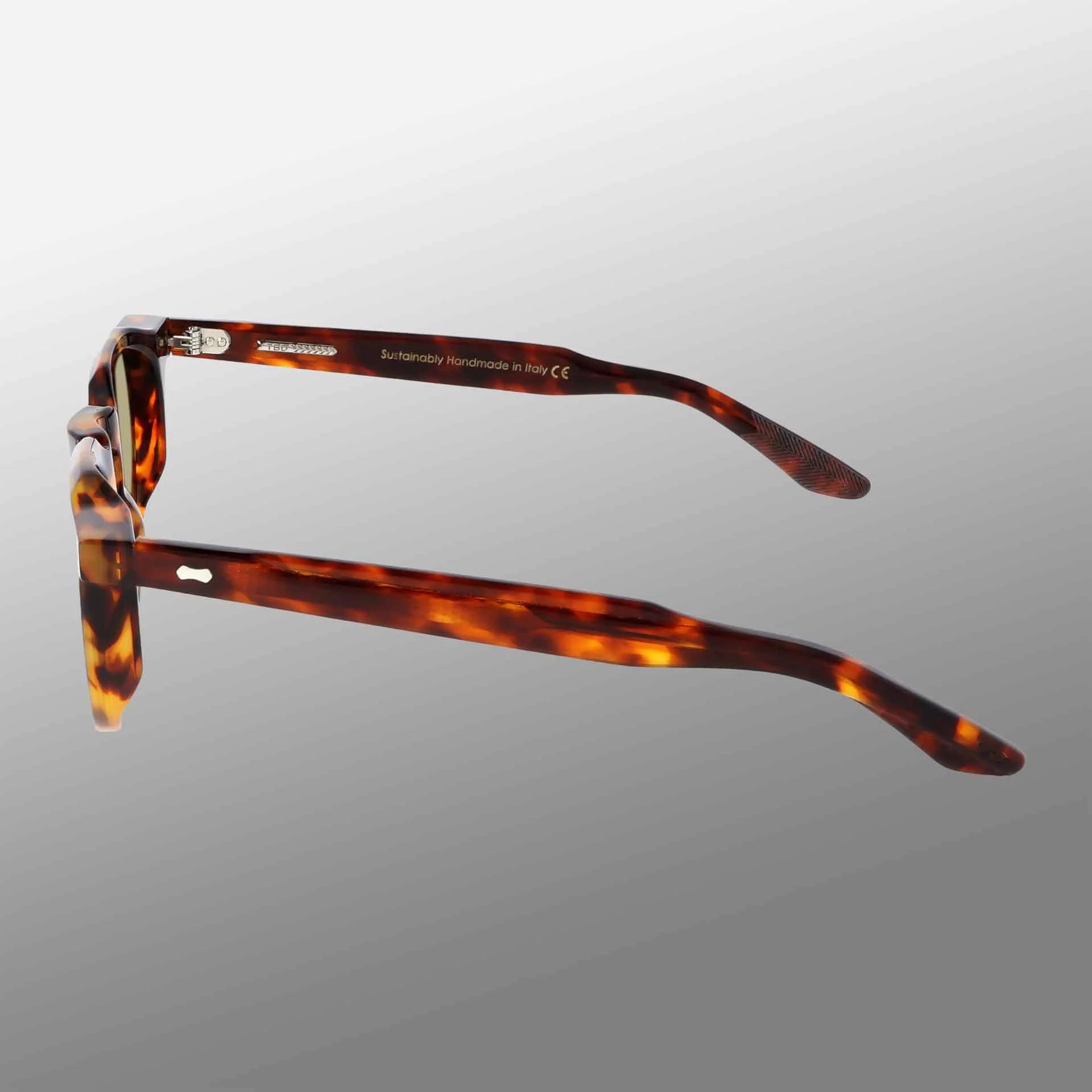 A pair of TBD Eyewear Hem Eco Spotted Havana | Mirror Green eyeglasses are shown from the back, displaying dark brown and amber-mottled arms with “Handmade in Italy” on the left arm, against a smooth gray gradient background. , available at Richard Gelding Mayfair