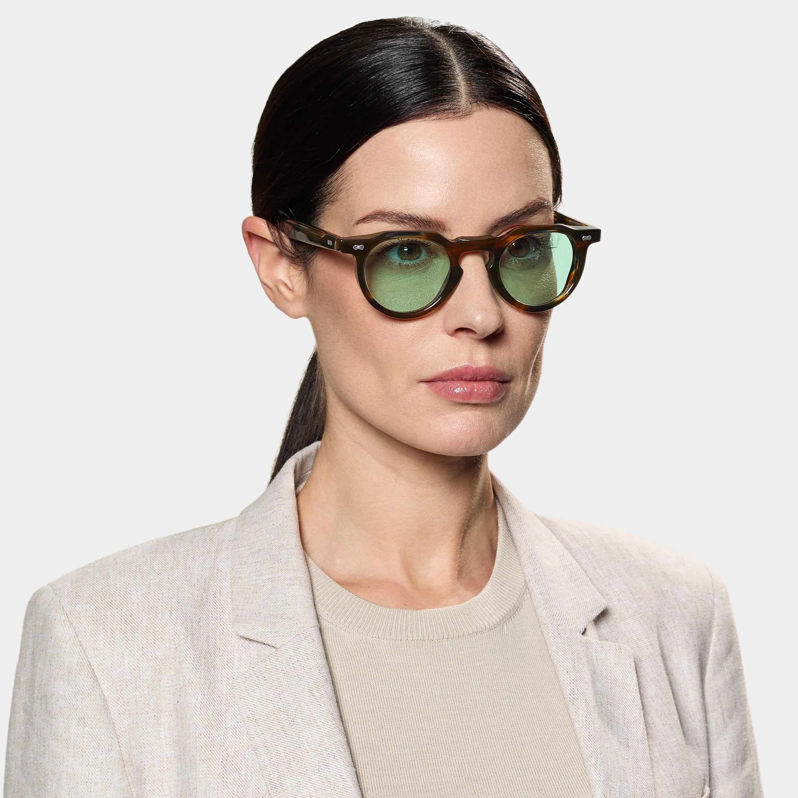 A woman with straight dark hair pulled back wears TBD Eyewear’s Hem River | Light Green round sunglasses, a light beige blazer, and a beige top as she faces forward with a neutral expression against a plain, light gray background. , available at Richard Gelding Mayfair