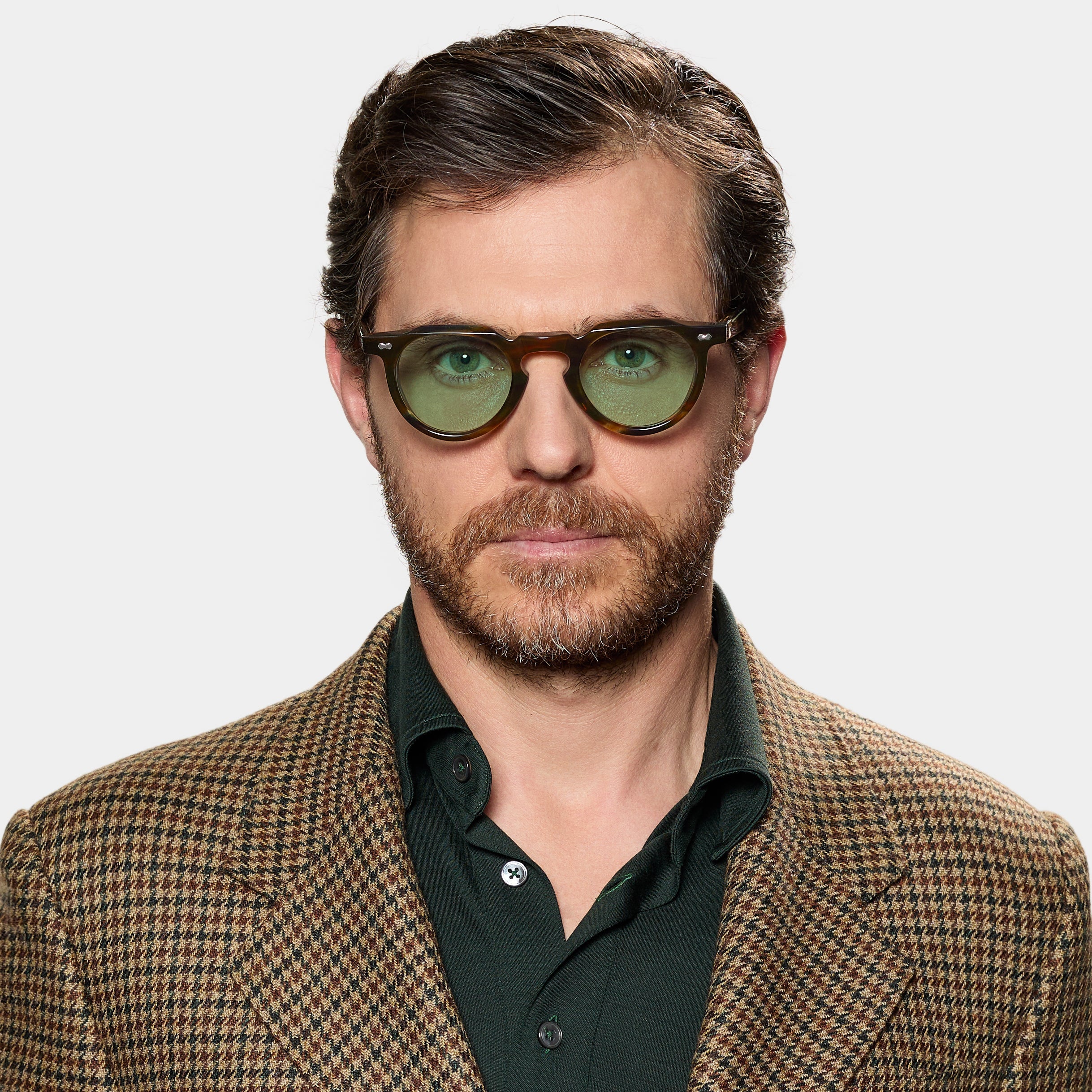 A man with short brown hair and a beard wears TBD Eyewear's Hem River | Light Green glasses. He looks serious in a brown houndstooth blazer over a dark green shirt, standing against a plain white background. , available at Richard Gelding Mayfair