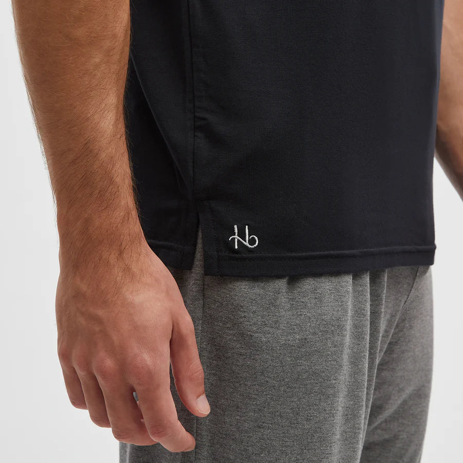 A close-up of a person’s left arm and torso wearing a Homebody Black Modal Classic Short Sleeve T-Shirt with a small white hb logo near the hem, paired with gray sweatpants. Their relaxed arm rests by their side against a plain white background. , available at Richard Gelding Mayfair