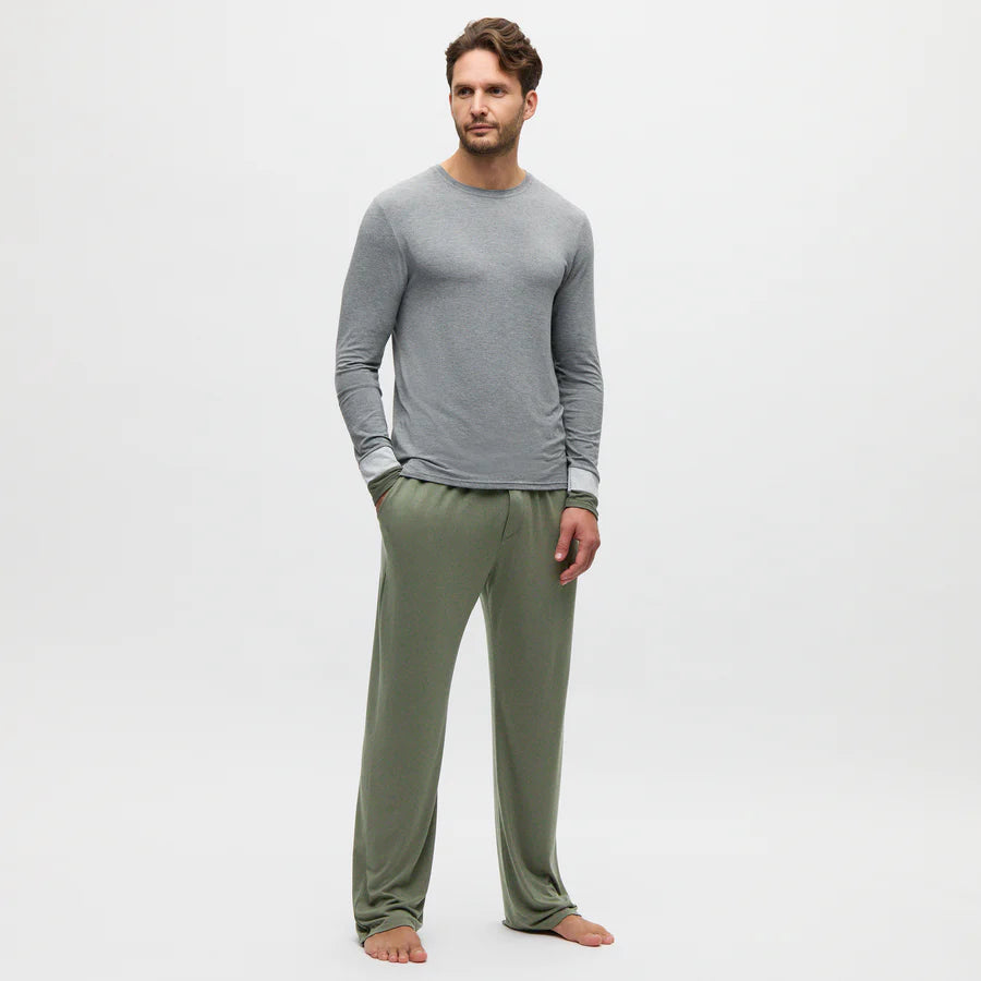 A man stands barefoot against a plain white background, wearing a light gray long-sleeve shirt and Homebody’s Charred Green Modal Duo Trousers. The trousers are loose-fitting, made from breathable fabric. He faces slightly left with a neutral expression, left hand in pocket. , available at Richard Gelding Mayfair