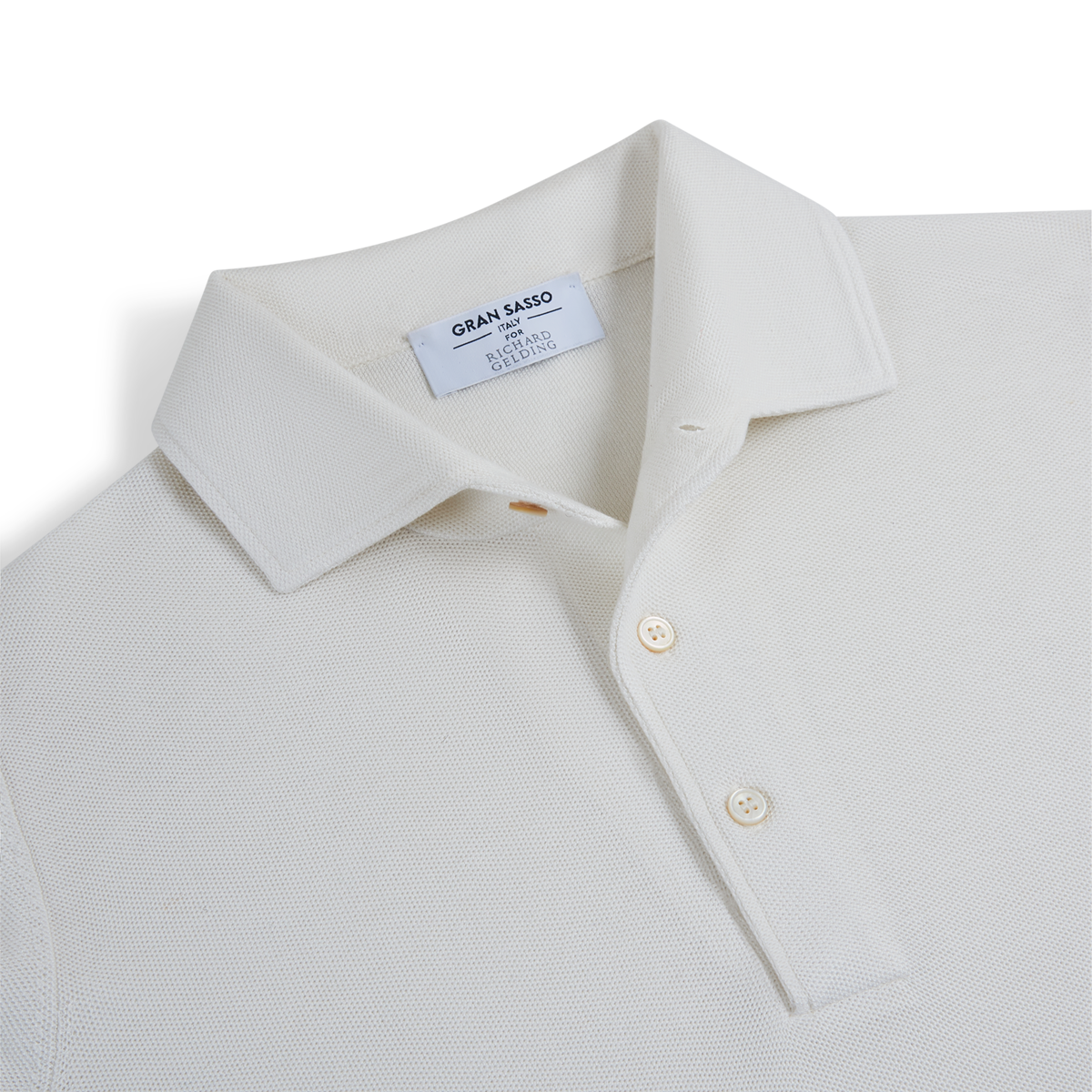 A close-up of the Gran Sasso White Silk Pique Polo shows its textured fabric, classic collar, and two cream buttons. The inside label displays “Gran Sasso,” “Made in Italy,” and notes the silk pique material. , available at Richard Gelding Mayfair