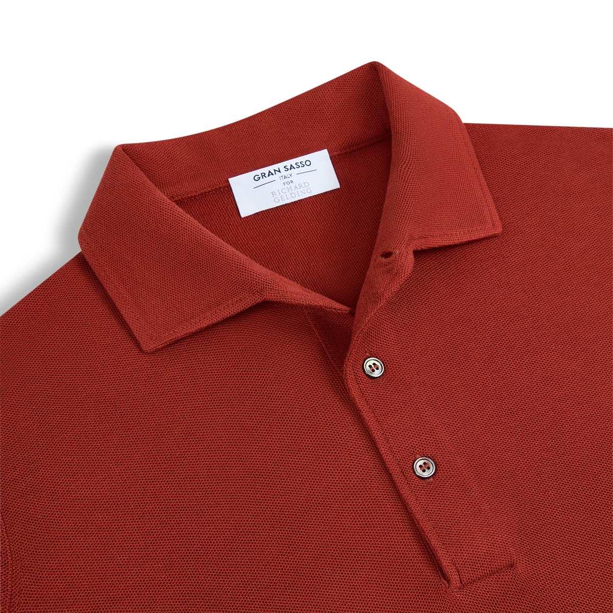 A close-up of the Rust Red Silk Pique Polo by Gran Sasso, featuring a textured knit, classic collar, two beige buttons, and an inner white “Gran Sasso” label, neatly laid flat on a white background. , available at Richard Gelding Mayfair