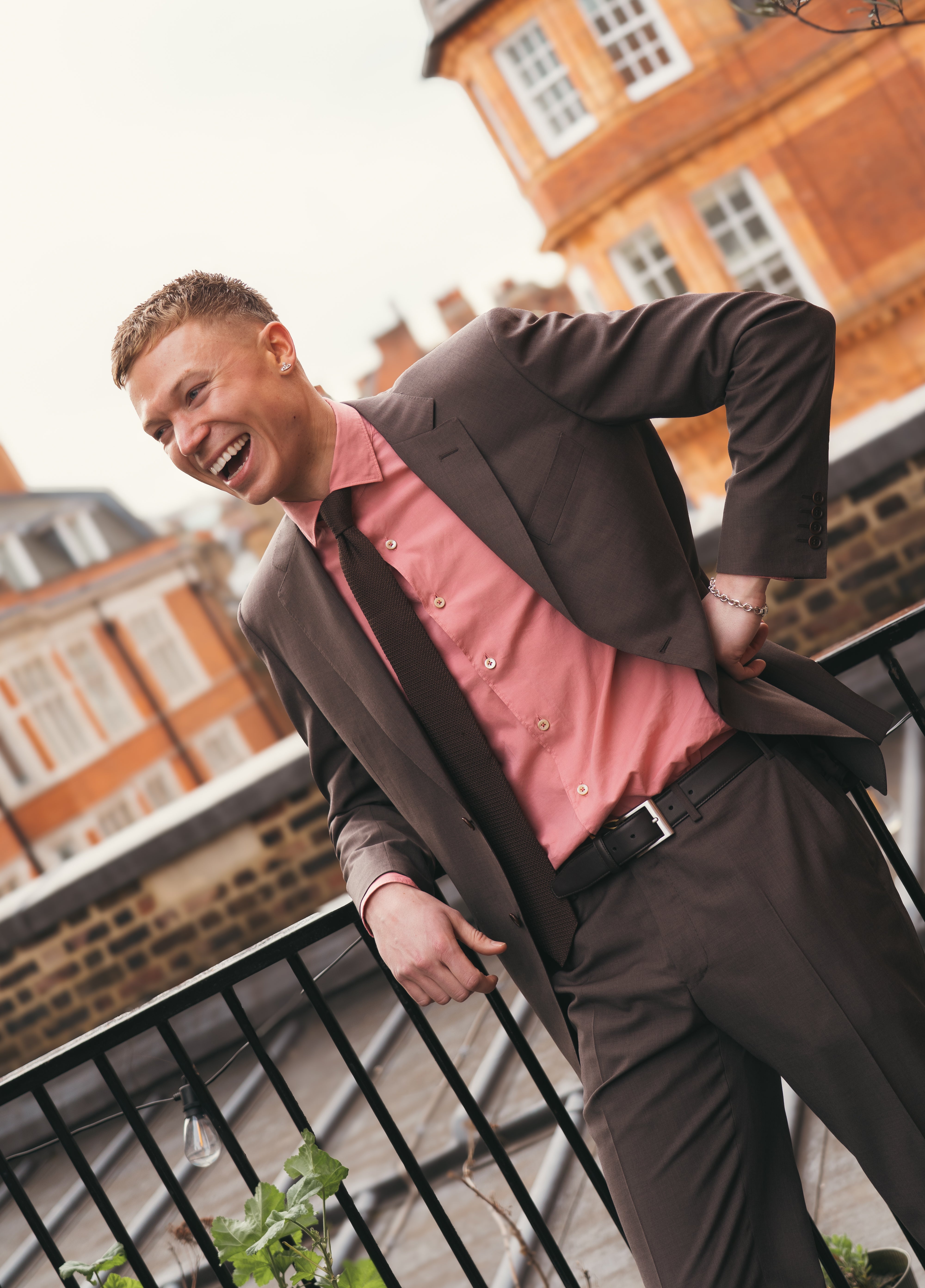 A cheerful man in a Canali Dark Brown Tropical Wool Suit and pink shirt leans on a black railing outdoors, red-brick buildings and windows softly blurred behind him, one hand on his hip and the other resting on the railing. , available at Richard Gelding Mayfair