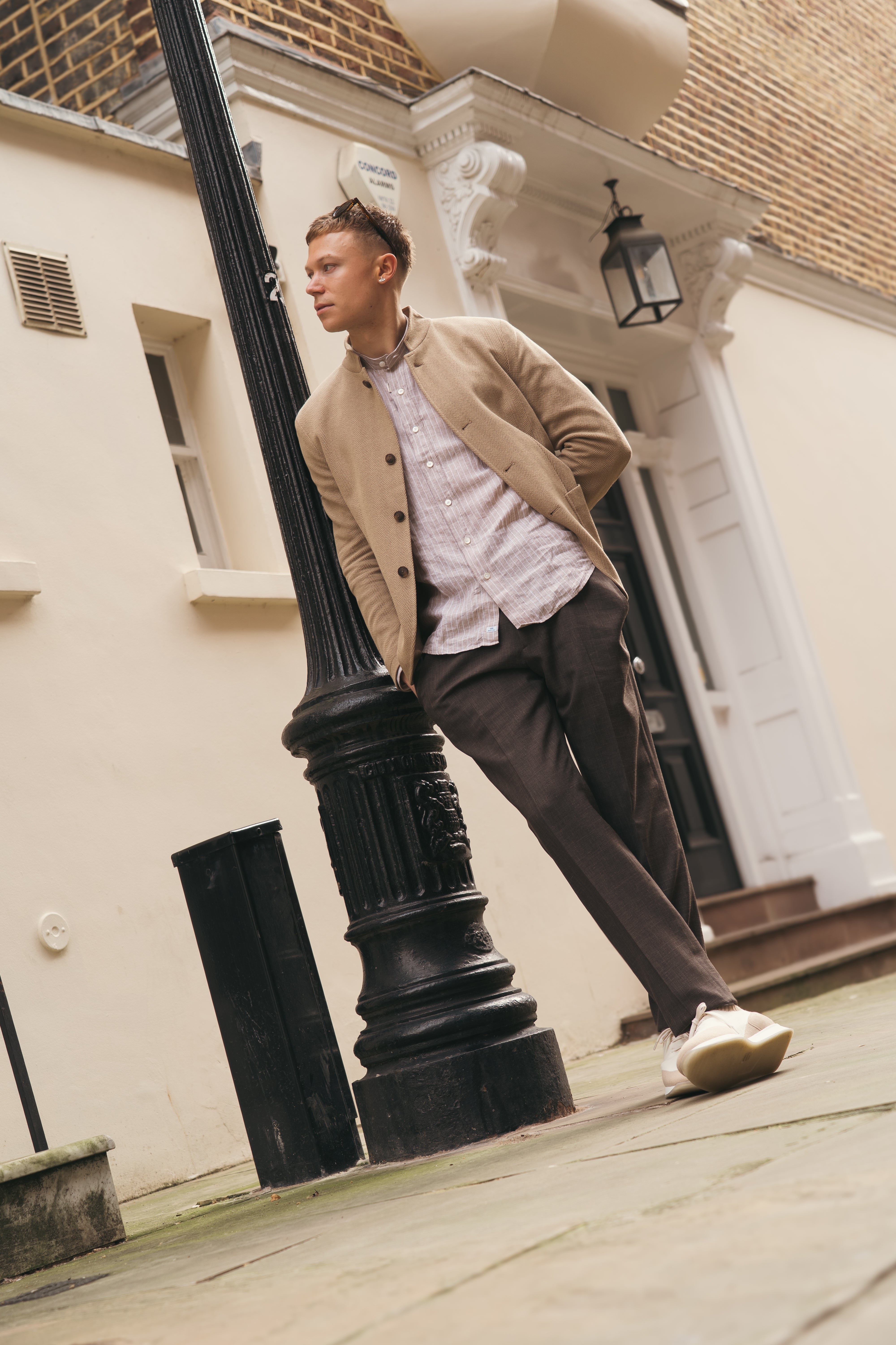 A man in a beige jacket, dark trousers, and an Alessandro Gherardi Brown Stripe Linen Mandarin Collar Shirt leans against a black lamppost on a stone-paved street before cream buildings with white trim and steps, hands in his pockets. , available at Richard Gelding Mayfair