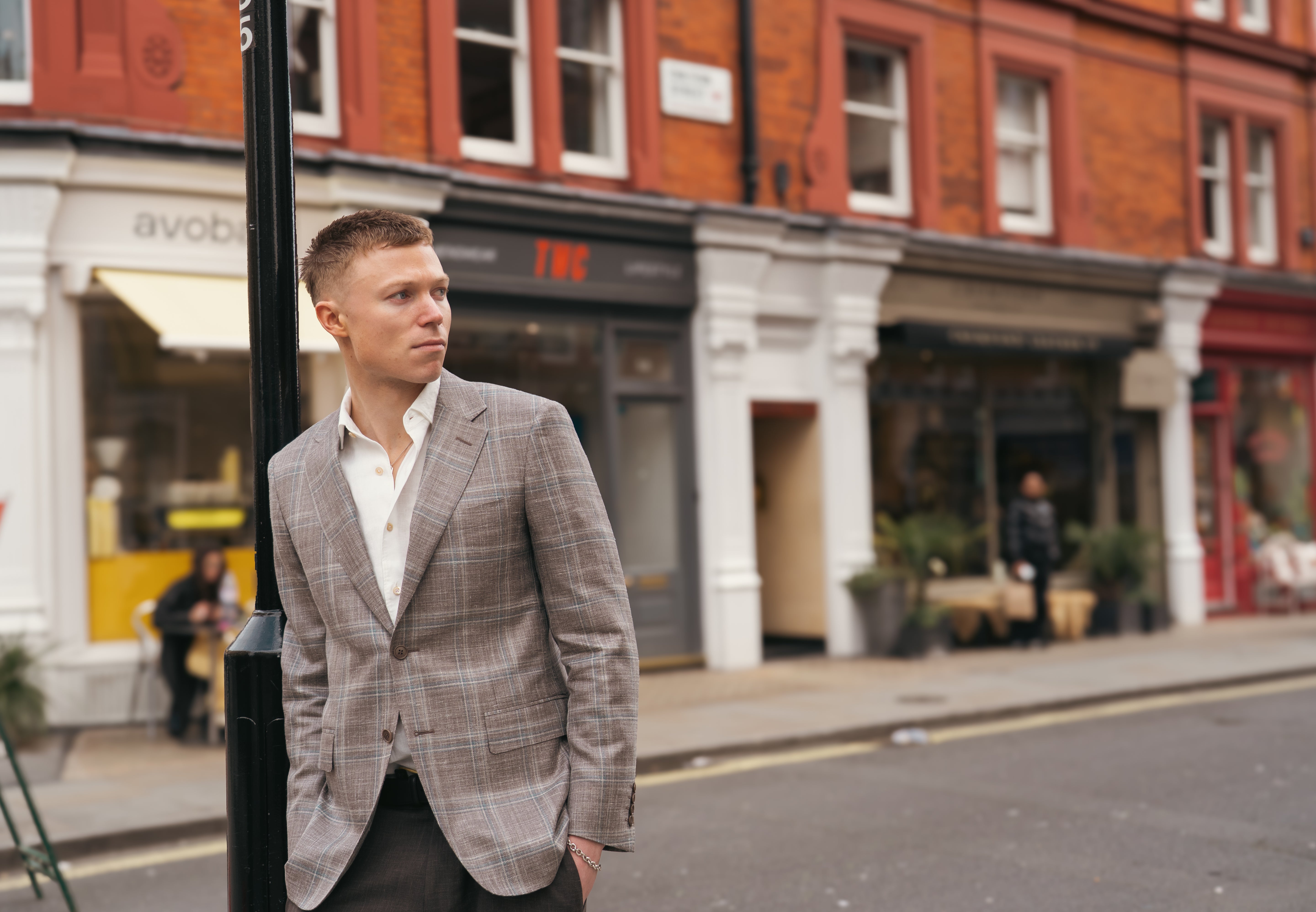 A man wearing the Canali Stone Teal Wool/Silk/Linen Windowpane Check Blazer and a white shirt stands on a city street, leaning against a black pole. Red-brick buildings with white and grey shopfronts form the soft-focused urban backdrop. , available at Richard Gelding Mayfair