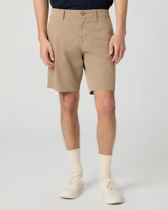 A person stands against a plain background wearing Paige Beige Farro Phillips Cotton Stretch Shorts, a dark shirt tucked in, white crew socks, and white sneakers. The image is cropped at the chest, hands relaxed at the sides. , available at Richard Gelding Mayfair