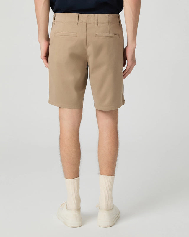 A person stands facing away from the camera in Paige Beige Farro Phillips Cotton Stretch Shorts with belt loops and two back welt pockets, paired with white crew socks and sneakers. The background is plain and light-colored. , available at Richard Gelding Mayfair
