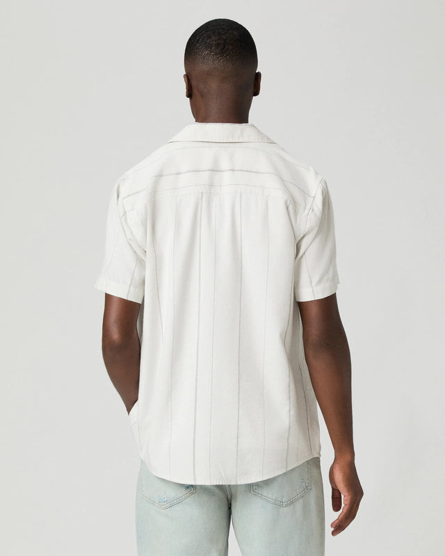 A man faces away from the camera in Paige's Ivory Stripe Cotton Blend 'Jacobson' Camp Collar Shirt, featuring subtle vertical stripes and short sleeves, paired with light blue jeans. A light gray background emphasizes his relaxed pose. , available at Richard Gelding Mayfair