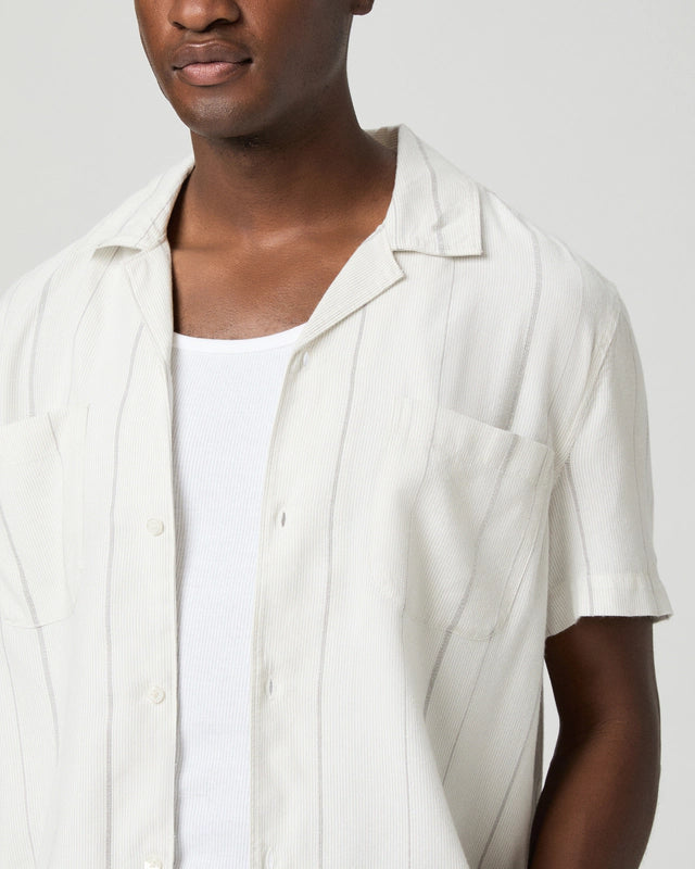 A person with medium skin tone wears the Paige Ivory Stripe Cotton Blend 'Jacobson' Camp Collar Shirt over a white ribbed tank, showing only their upper torso against a plain light gray background and a neutral expression. , available at Richard Gelding Mayfair