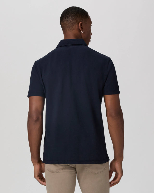 A man with short hair stands with his back to the camera, wearing the Paige Navy Pique Cotton Demille Short Sleeve Polo and light khaki pants, his arms relaxed at his sides against a plain light gray background. , available at Richard Gelding Mayfair