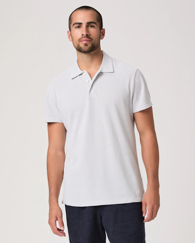 A man with short hair and a trimmed beard stands facing forward in the Paige Bristol Blue Demille Cotton Polo, a classic short-sleeve men's shirt, paired with dark pants. He has a neutral expression against an off-white background. , available at Richard Gelding Mayfair