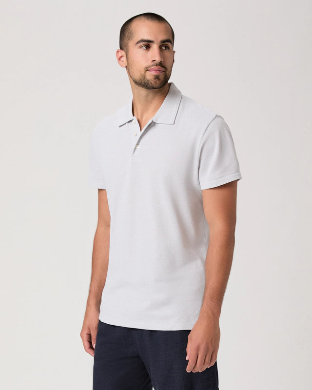 A man with a trimmed beard and short hair wears the Paige Bristol Blue Demille Cotton Polo with dark pants. He stands against a plain light background, looking slightly left with a neutral expression and relaxed posture. , available at Richard Gelding Mayfair