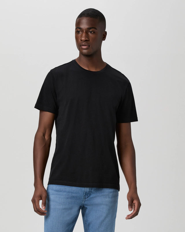 A man stands against a light gray background wearing the Paige Black Cotton Sorden Crew Neck T-Shirt and light blue jeans. He looks slightly to the side with a neutral expression, arms relaxed by his sides. , available at Richard Gelding Mayfair